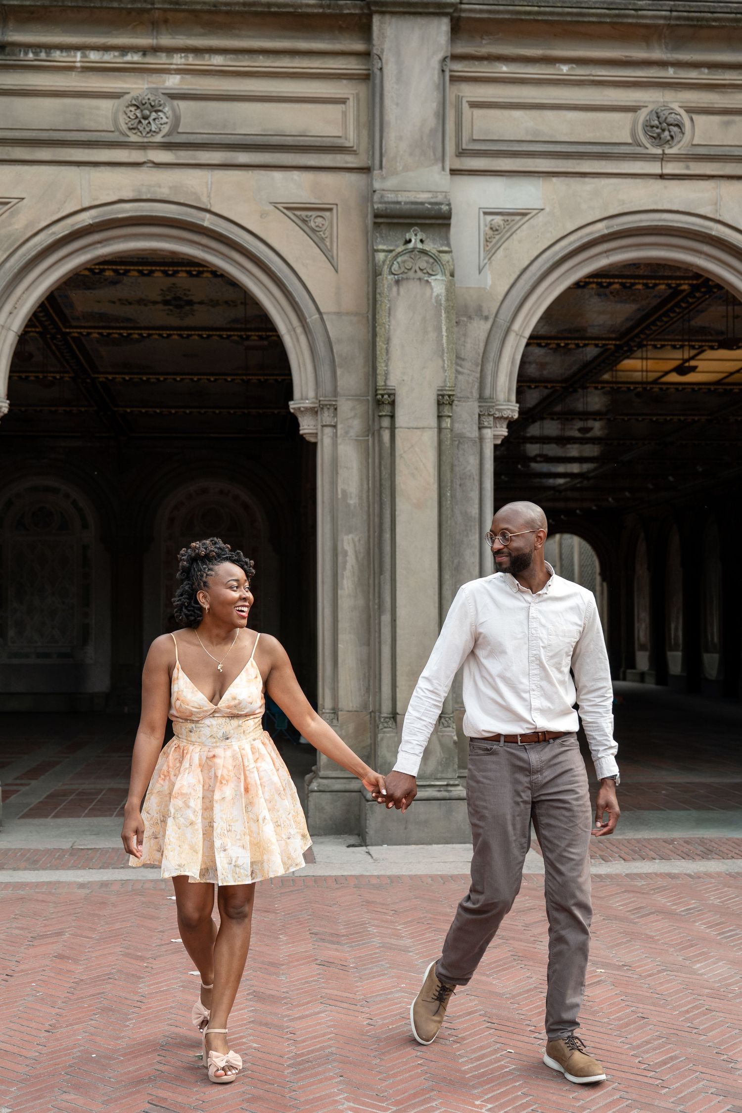 A couple walks hand in hand under ornate stone archways in an elegant architectural setting.