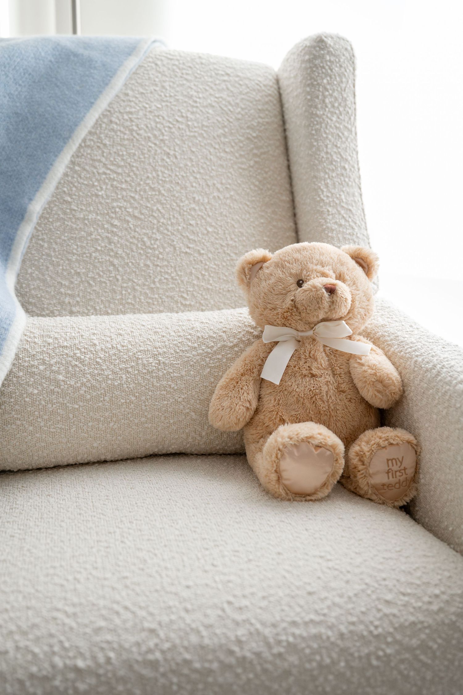 A soft beige teddy bear sits alone on a plush white couch.