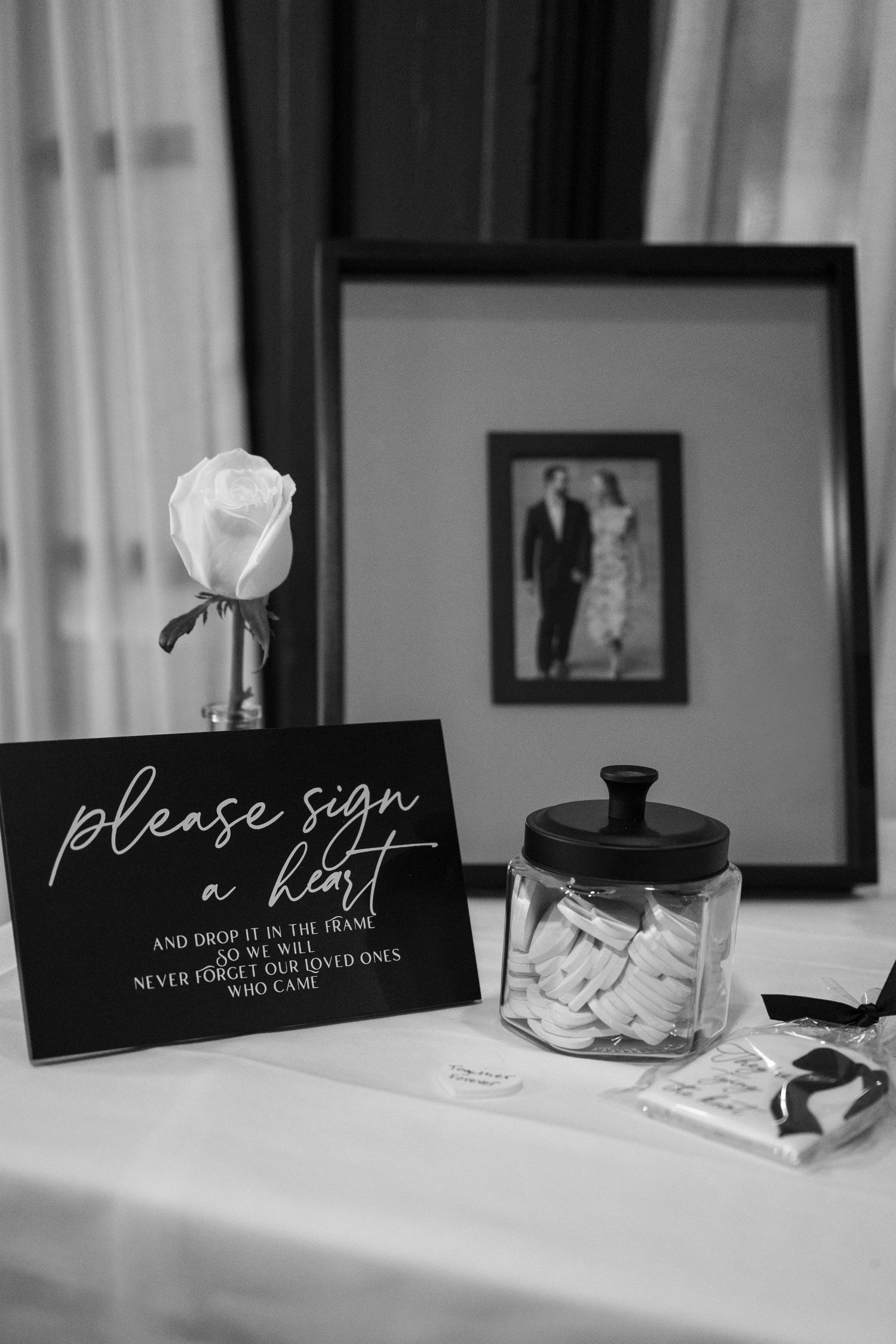 Black and white wedding detail shot of a guest book signing station with a white rose and glass jar on a table.