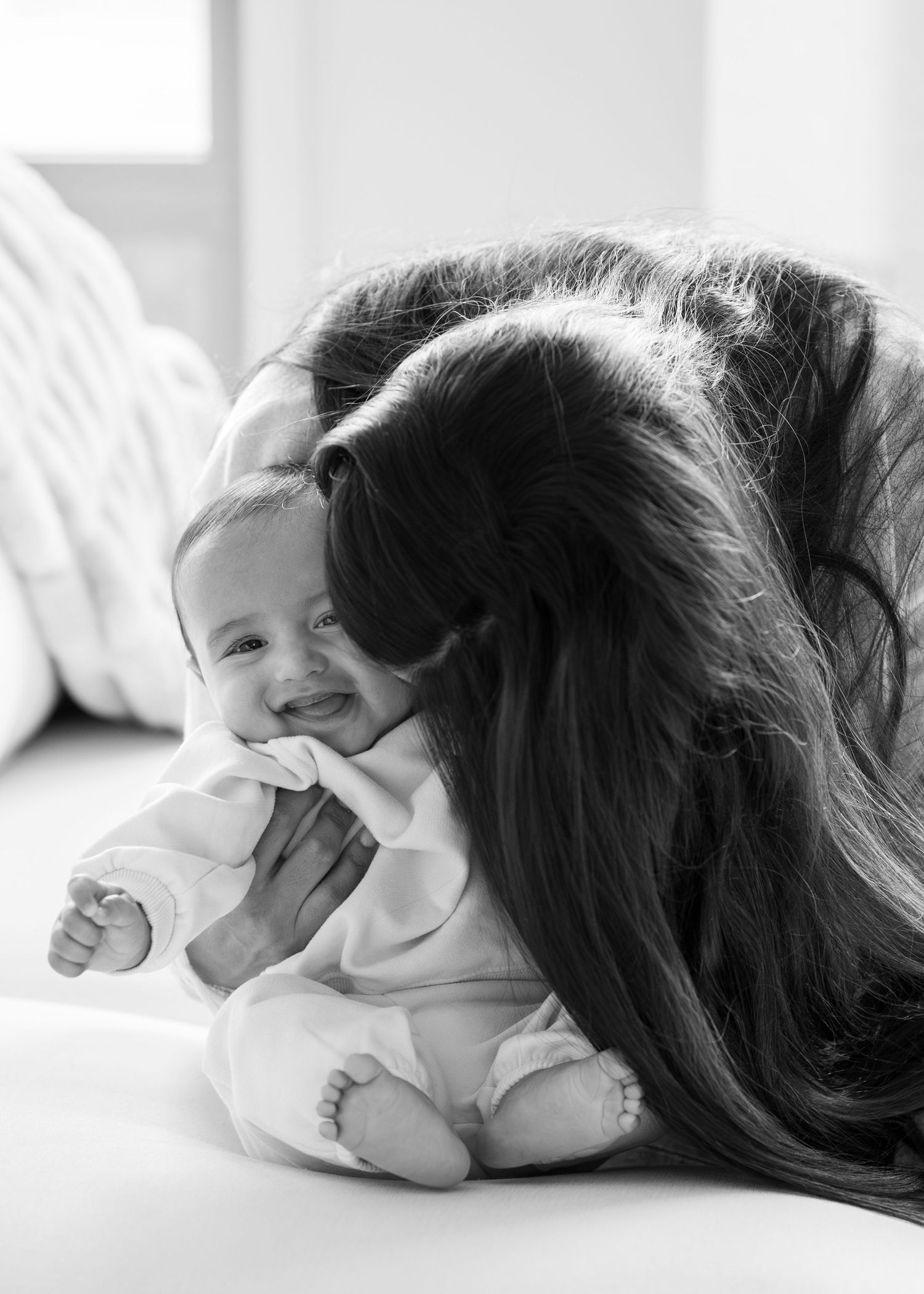 Black and white photo of a mother tenderly nuzzling her smiling newborn baby on a soft white surface.