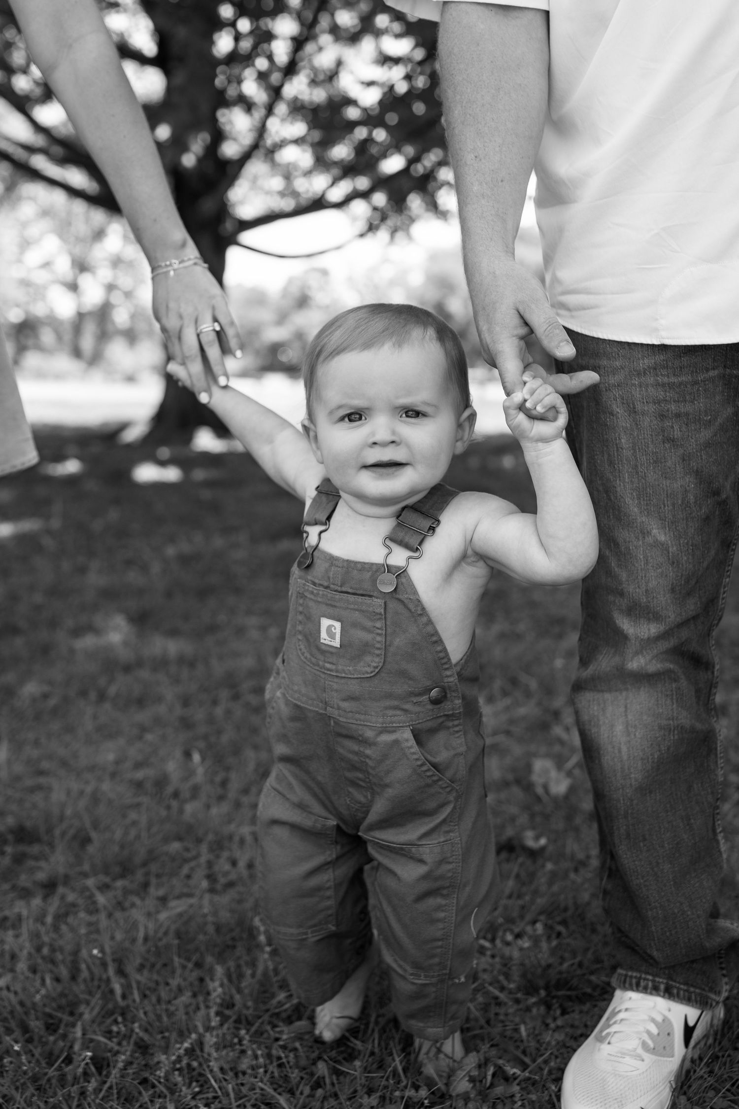 Black and white photo of a baby in overalls taking first steps while parents hold hands outdoors.