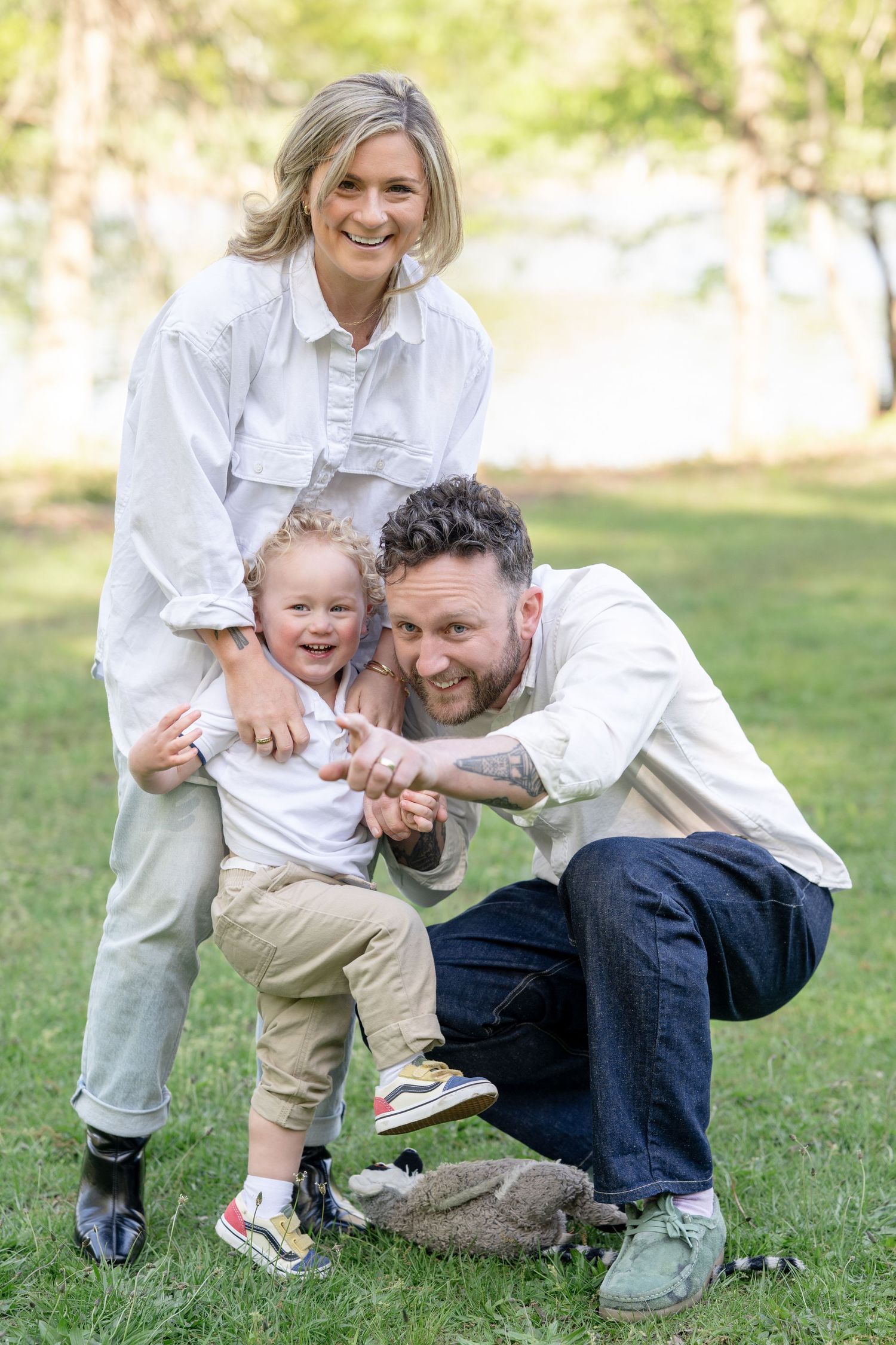 Family of three smiling outdoors in park, parents in white shirts with curly-haired toddler holding a stuffed animal.