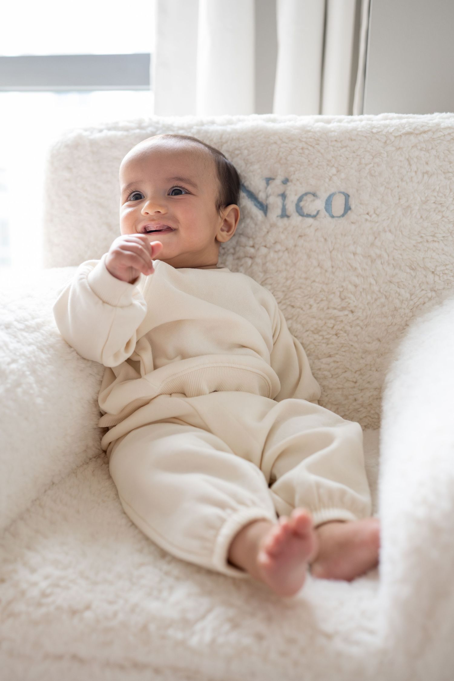 A baby wearing cream-colored clothing lies on a white couch while smiling and playing.