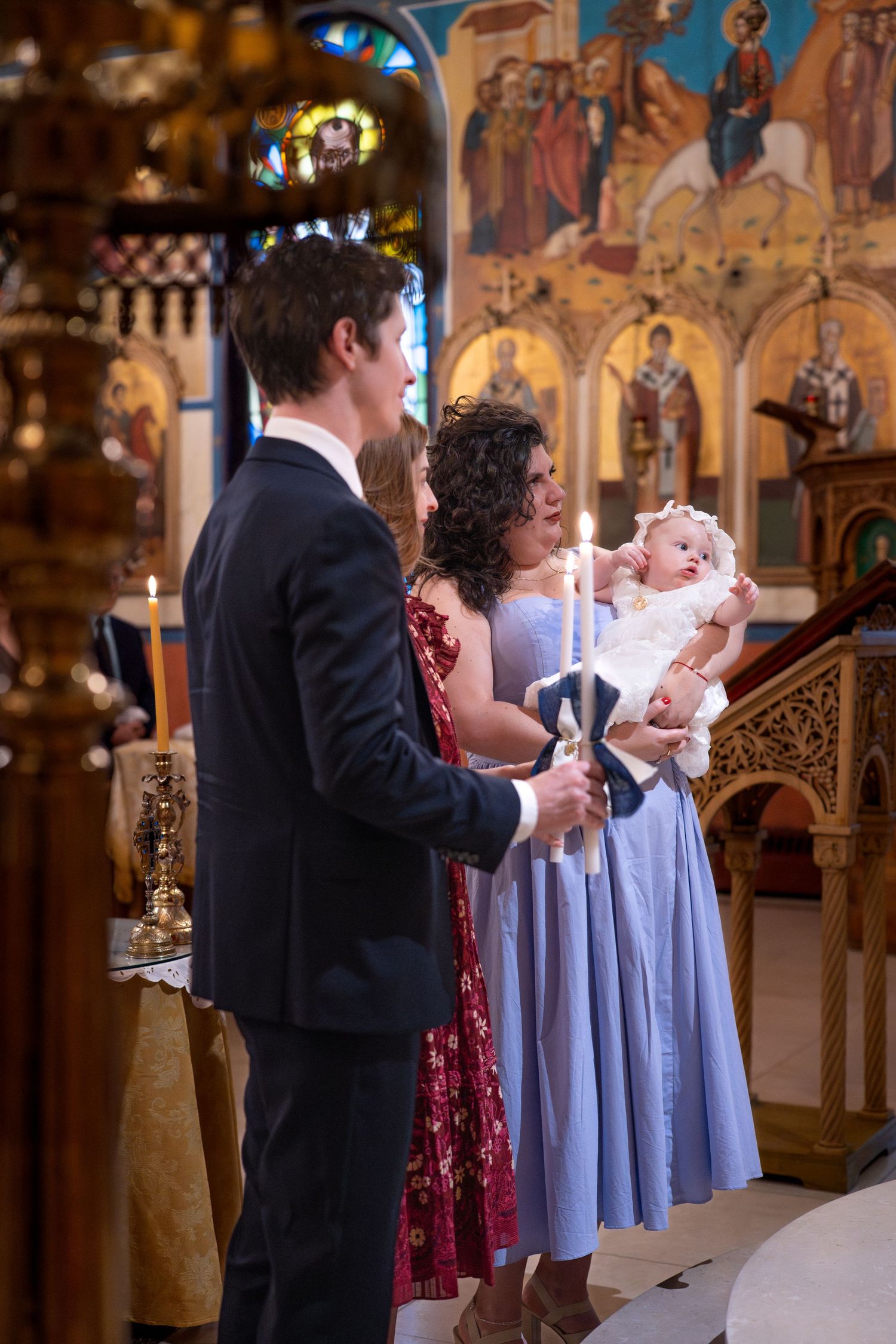 A baptism ceremony in progress with attendees standing in an Orthodox church with decorated walls.