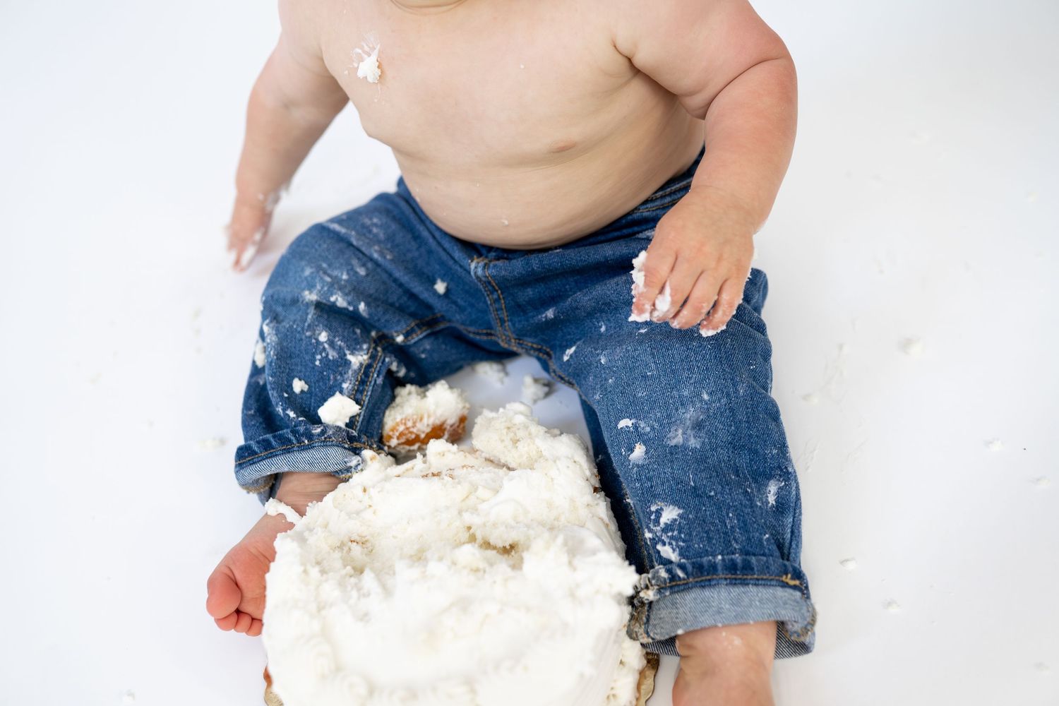 Overhead view of a baby in denim jeans smashing a white frosted cake during a first birthday cake smash.