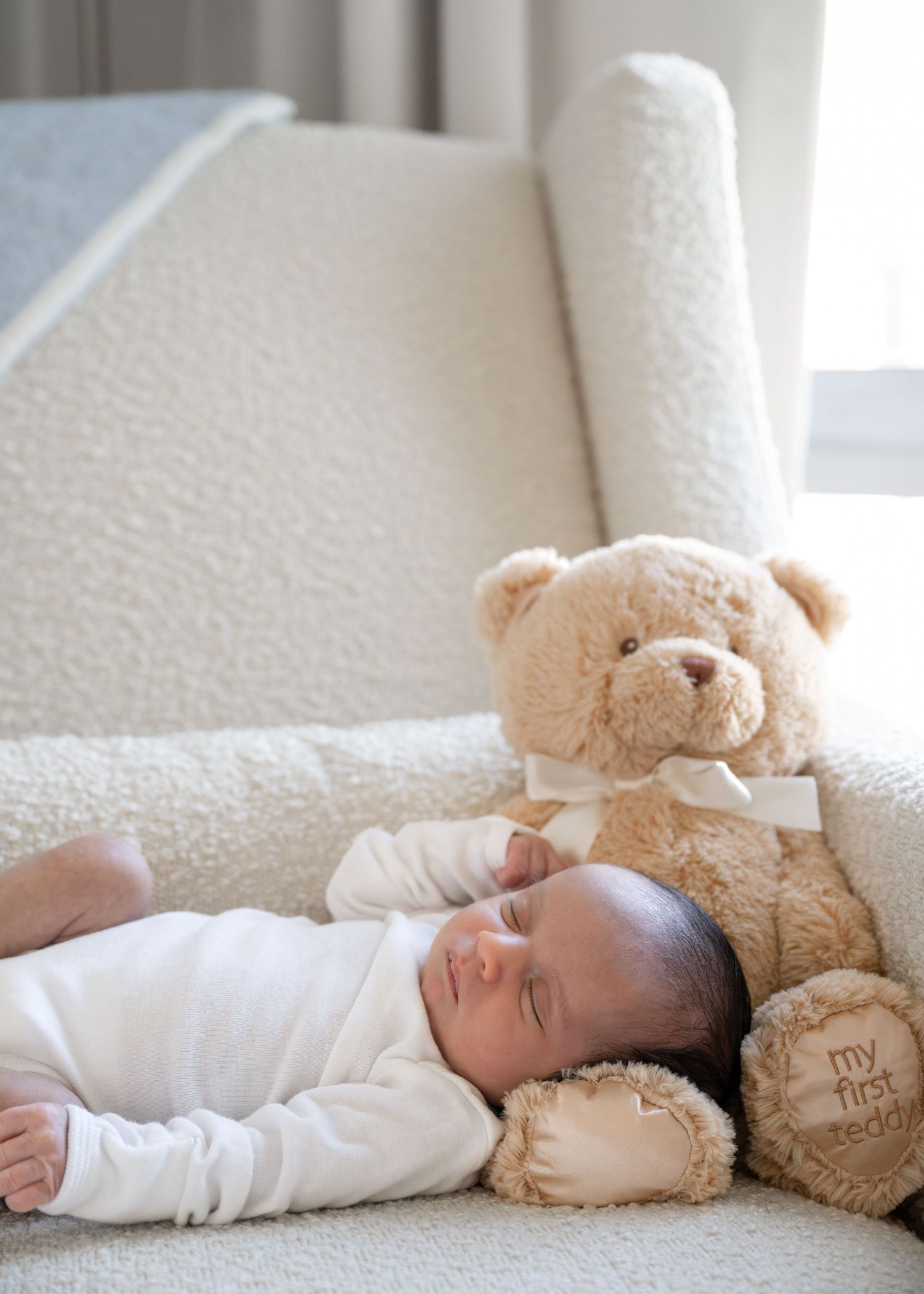 Sleeping newborn in white outfit cuddled next to plush teddy bear on white bedding.