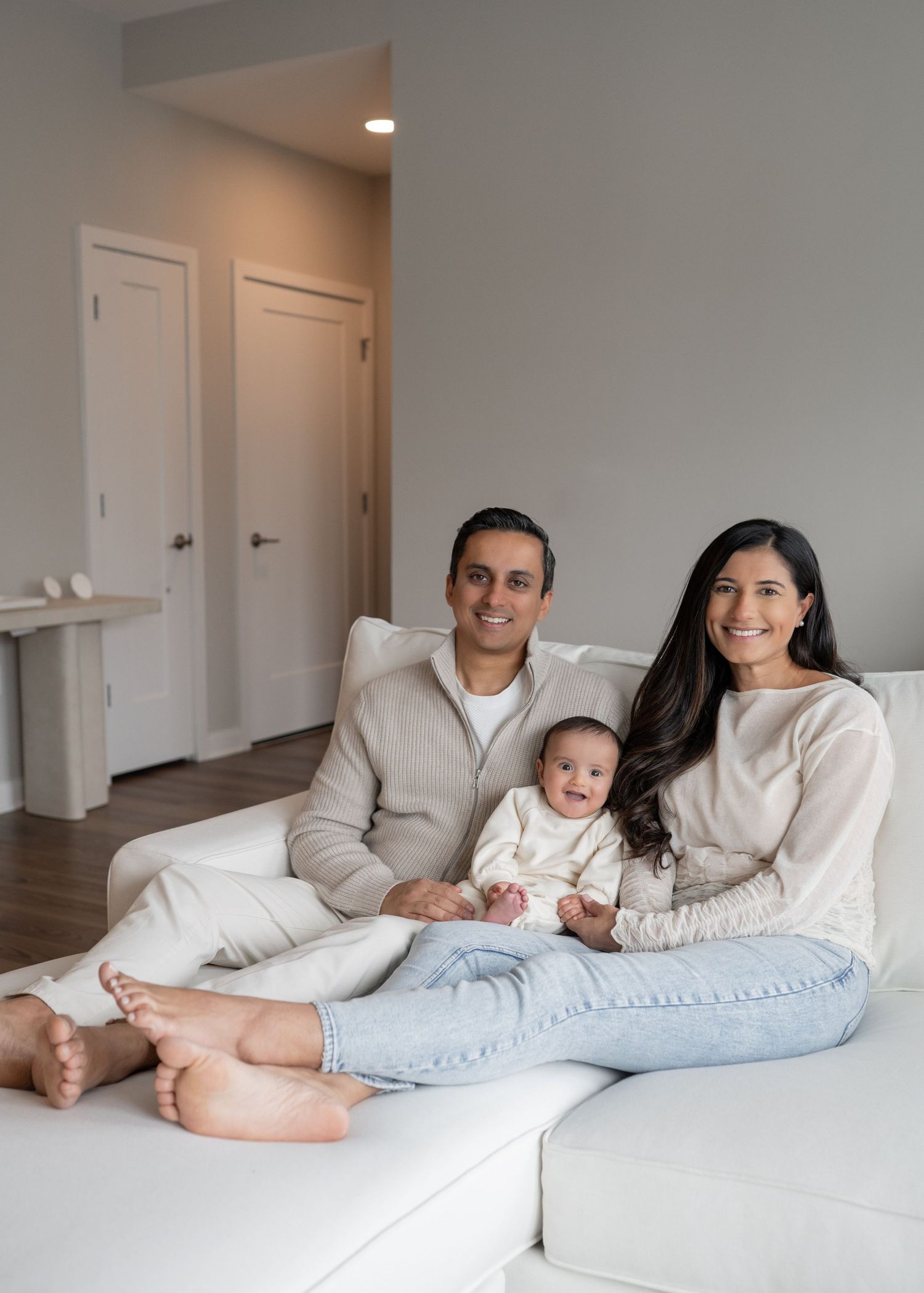 Smiling family with baby in white romper relaxing together on white sofa in modern home.