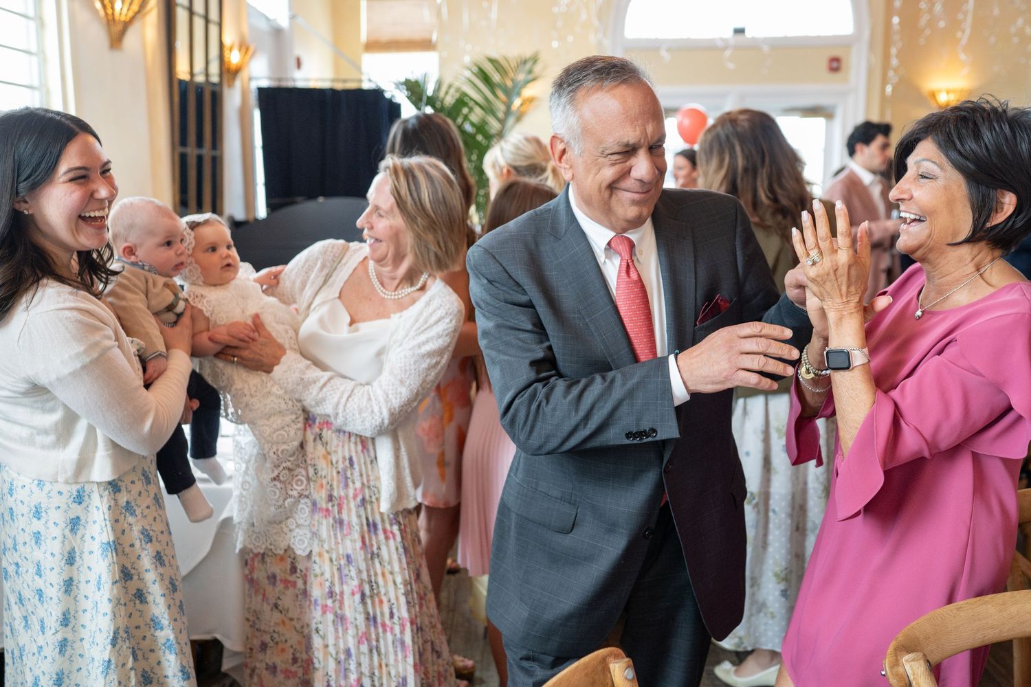 Guests laughing and celebrating at an indoor party event with a baby being held in the background.