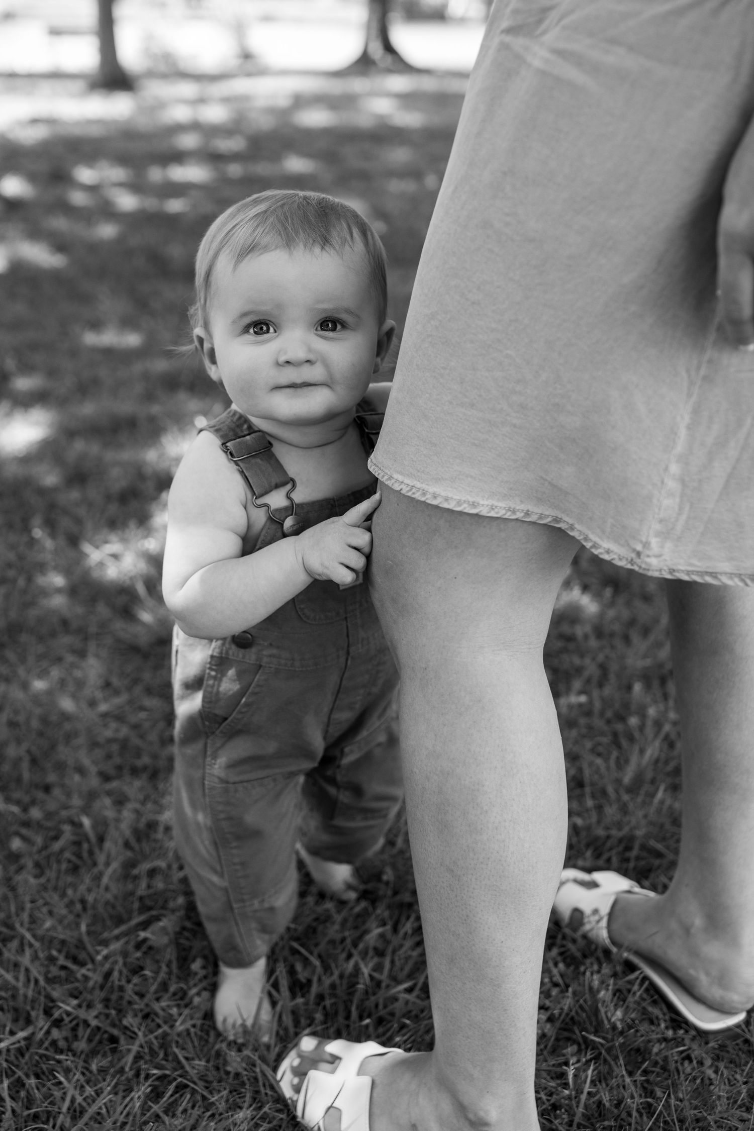 Adorable baby in overalls clinging to mothers leg while standing on grass in black and white photo.