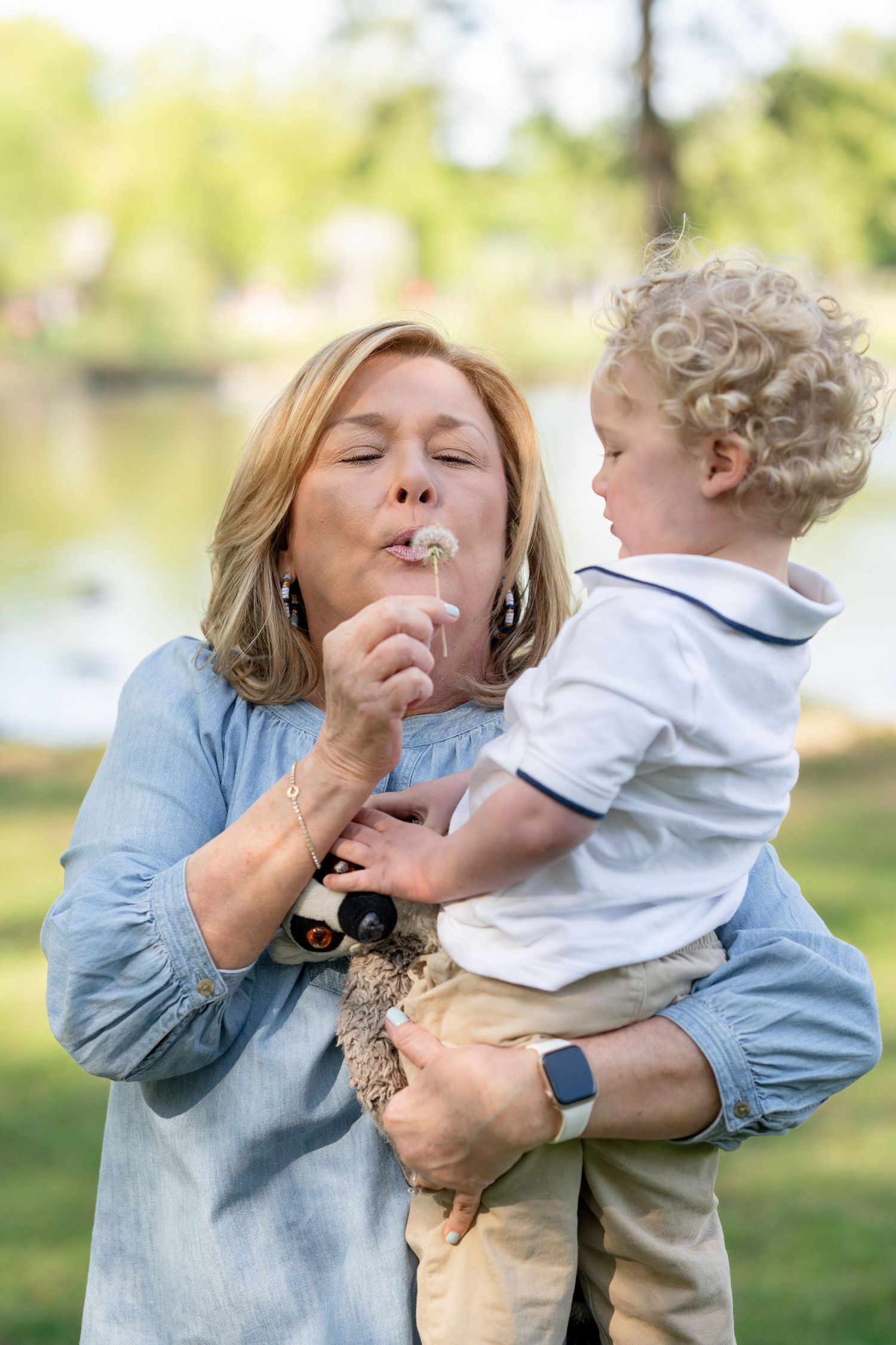 A grandmother blows a dandelion while holding her curly-haired toddler grandson in a sunny park.