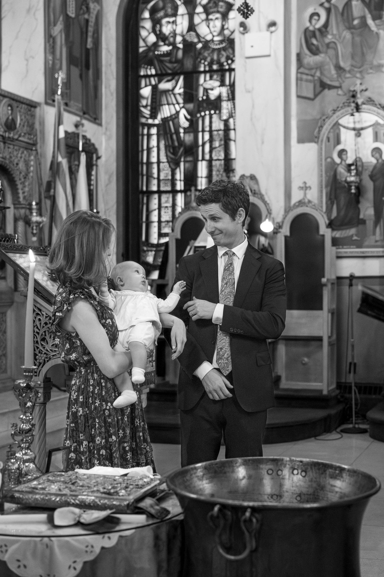 A christening ceremony takes place inside a church with ornate stained glass windows and religious artwork visible.