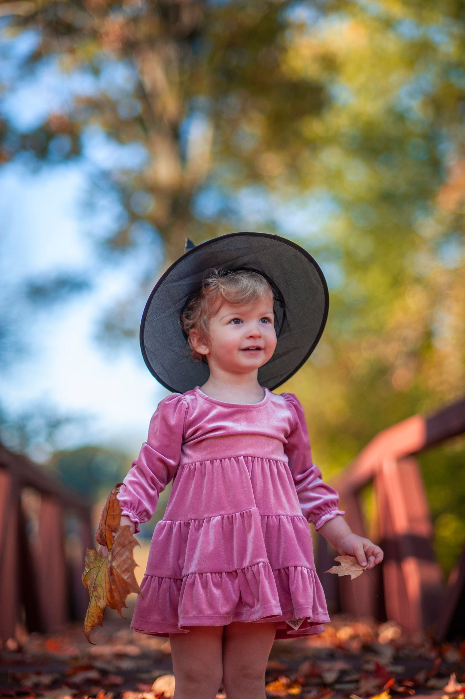 Child in pink dress and wide-brimmed hat plays in autumn leaves.