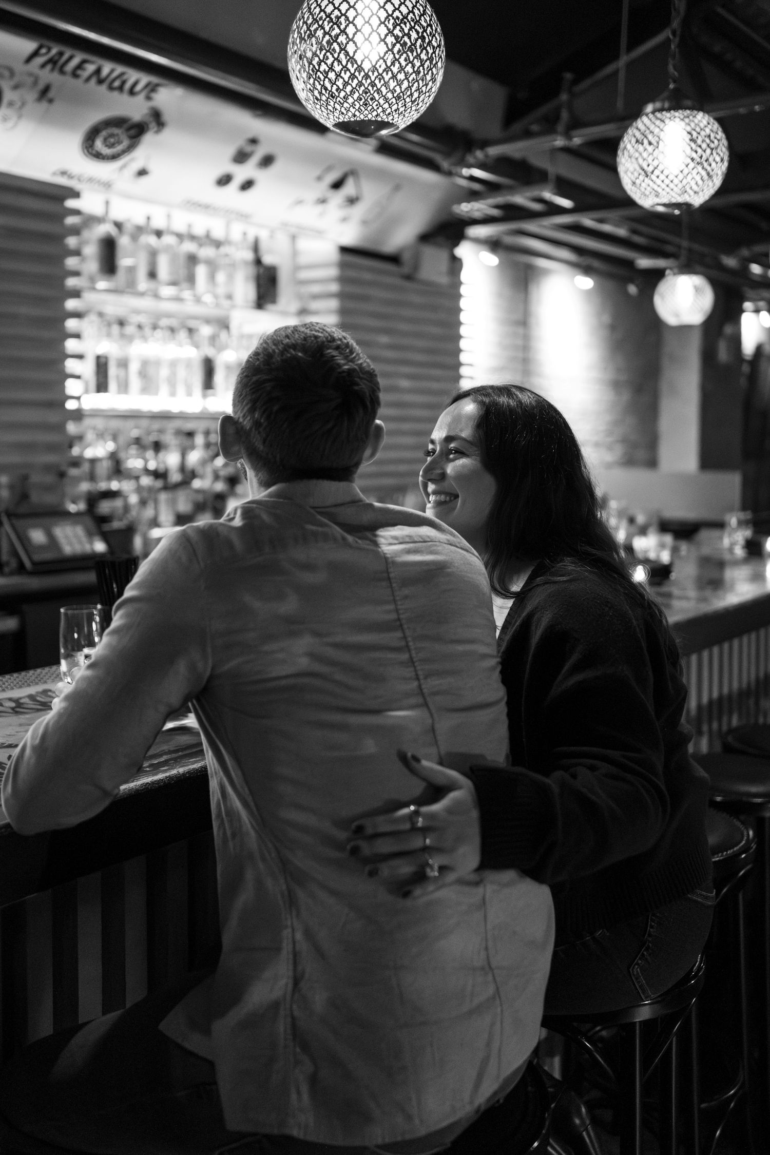 A couple sitting close together at a bar under crystal pendant lights in black and white.