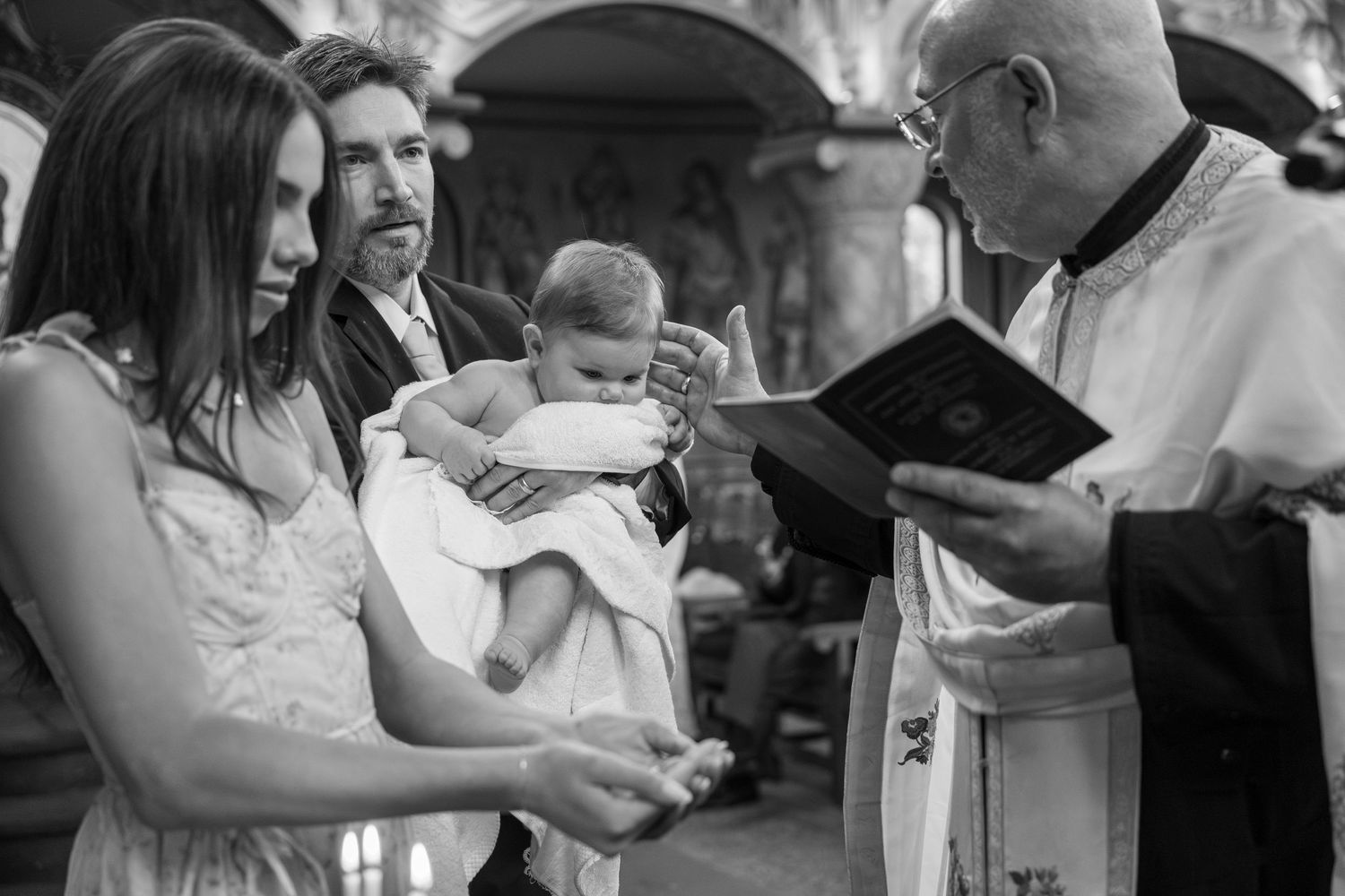 Black and white photo of a baby baptism ceremony with parents and priest reading from a religious book in a church.
