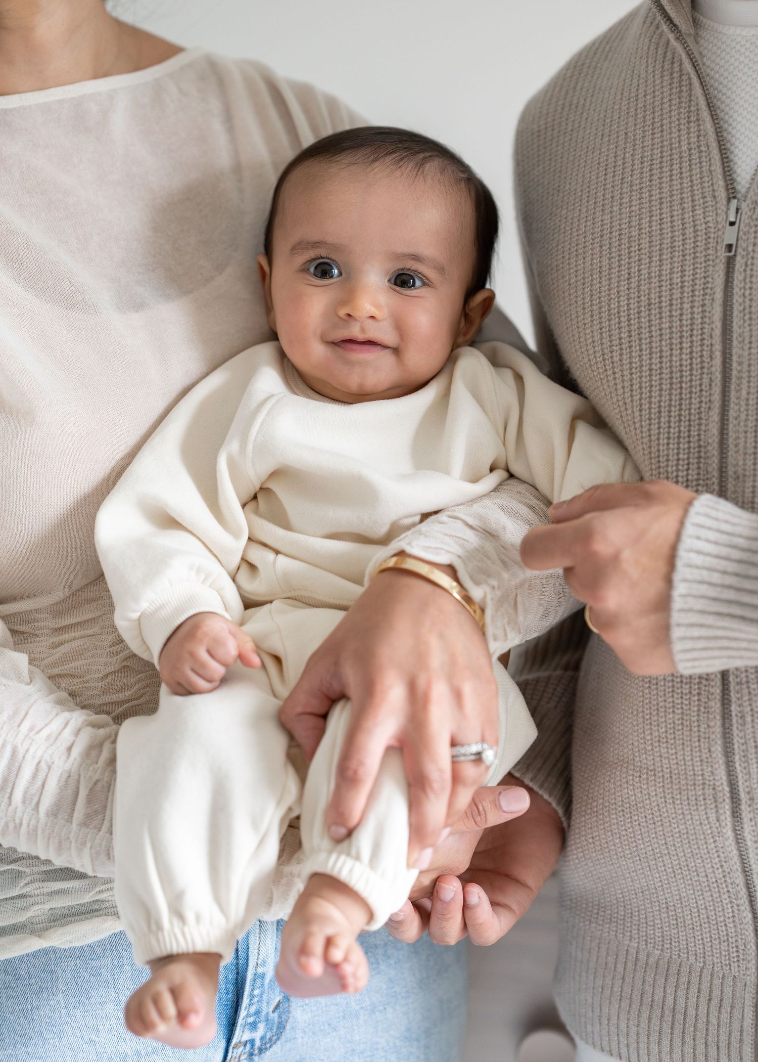 Adorable baby in cream outfit held by parents, smiling at camera.