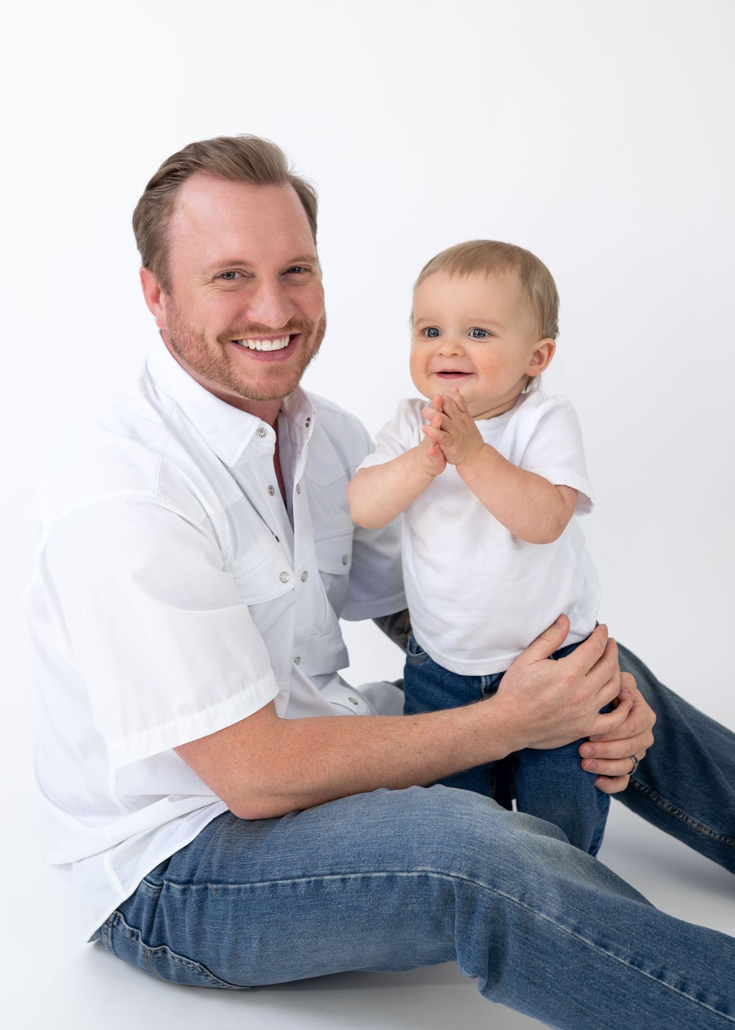 Smiling father sitting with happy baby clapping hands, both wearing white shirts and jeans.