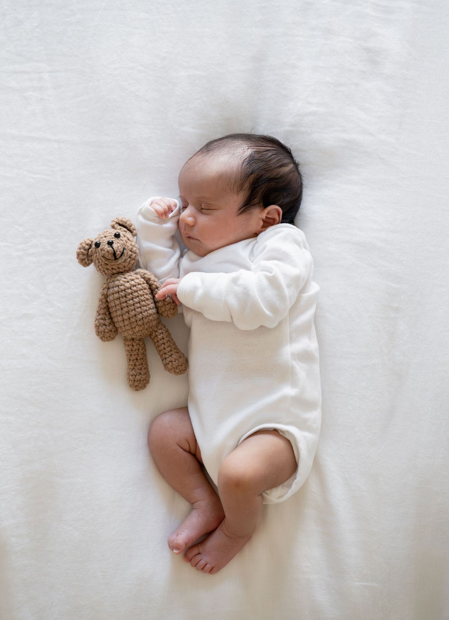Newborn in white onesie sleeping next to small brown knitted teddy bear.