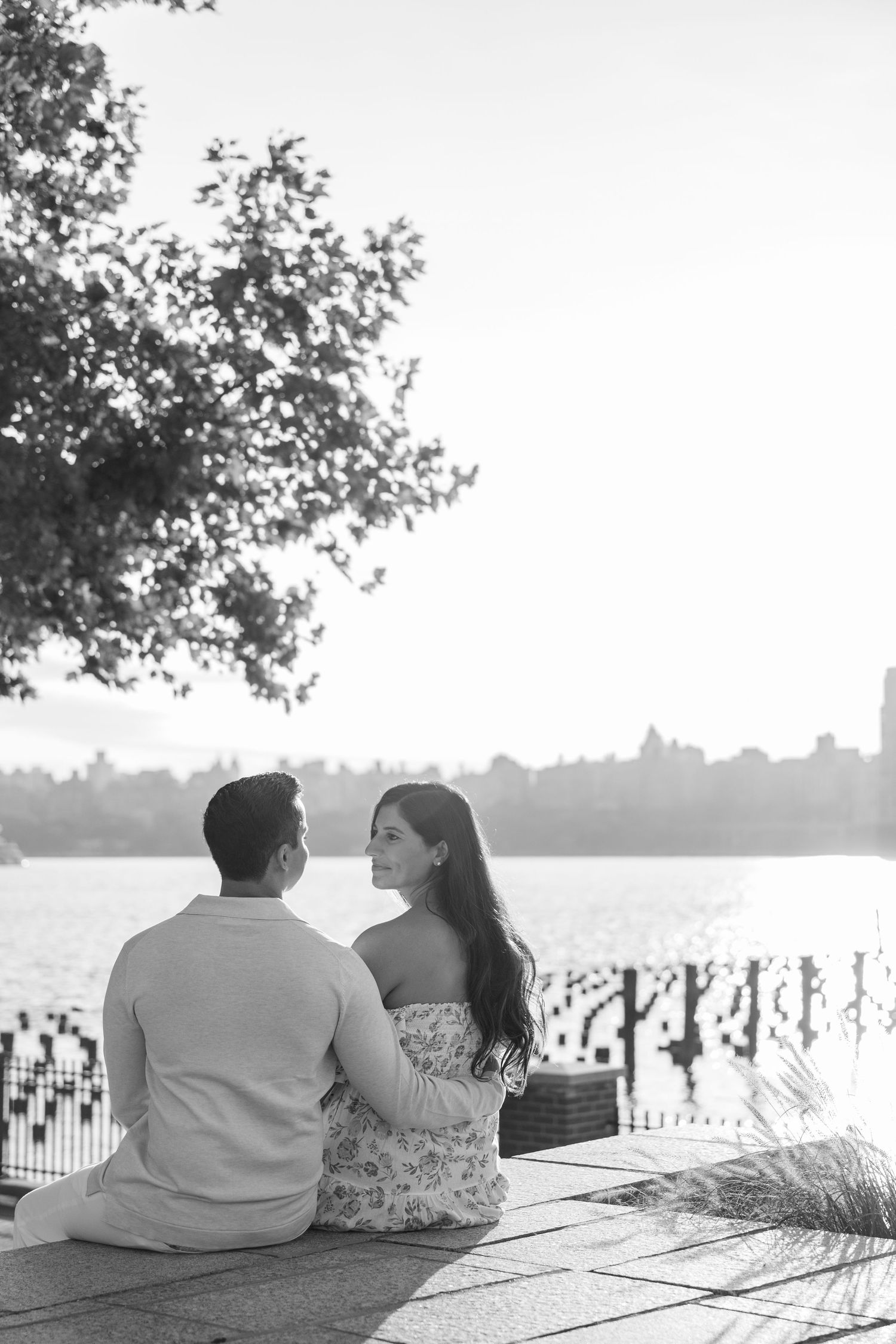 Couple sitting together on a bench overlooking a scenic waterfront with trees in the background.