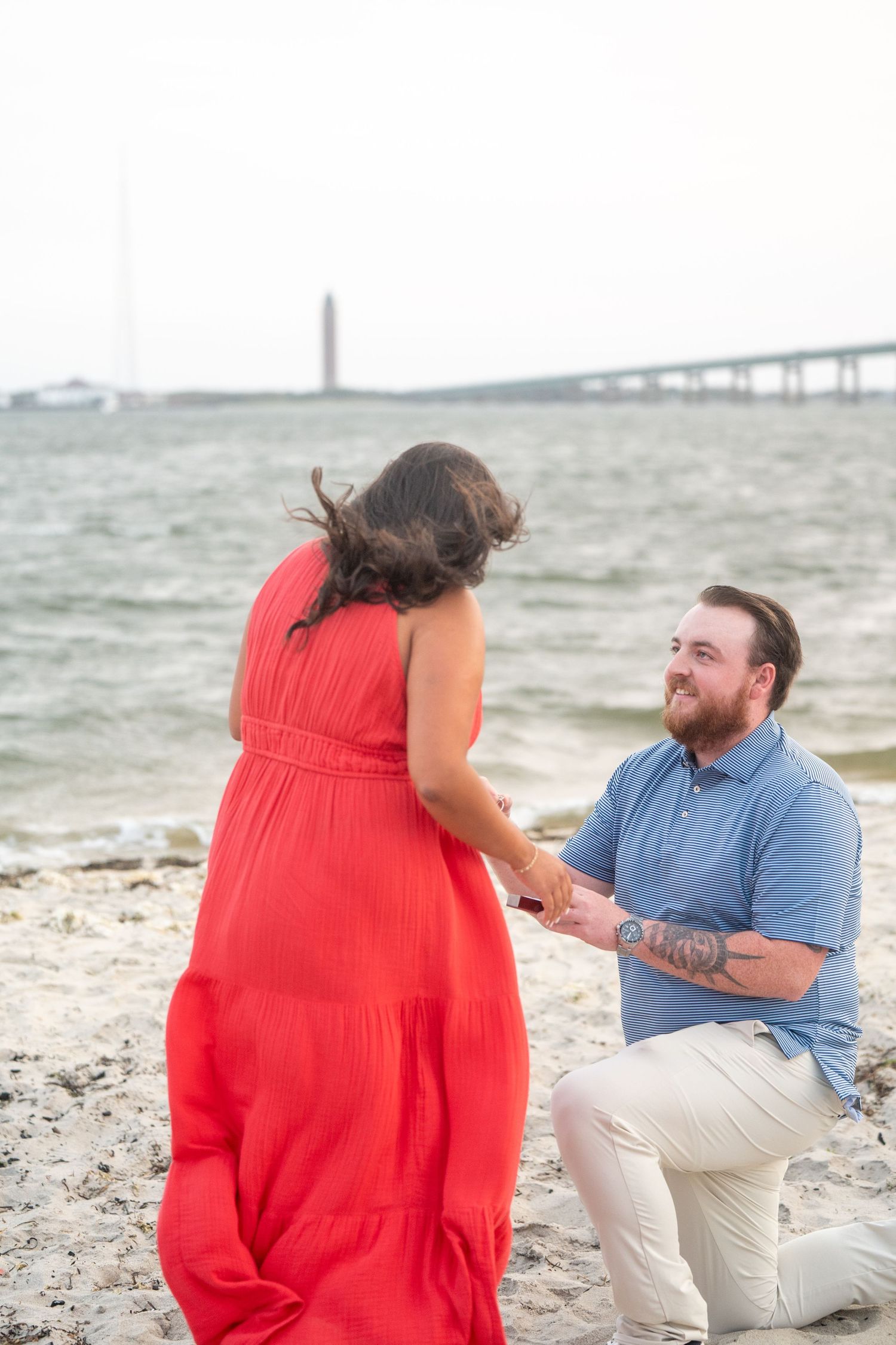 A romantic beach proposal scene with a bridge in the background as someone kneels on one knee in the sand.