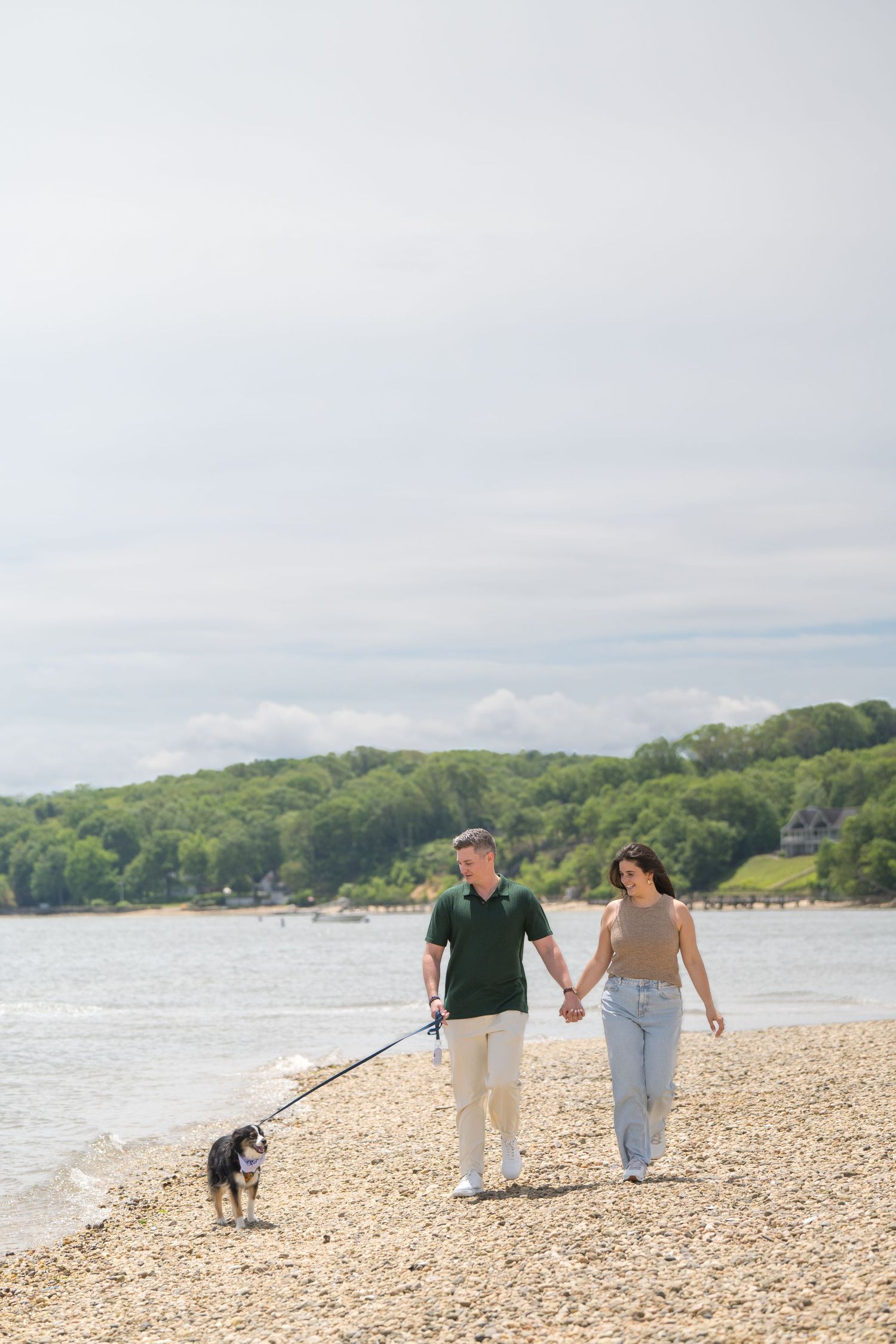 A couple walks their dog along a scenic rocky beach with trees and water in the background.