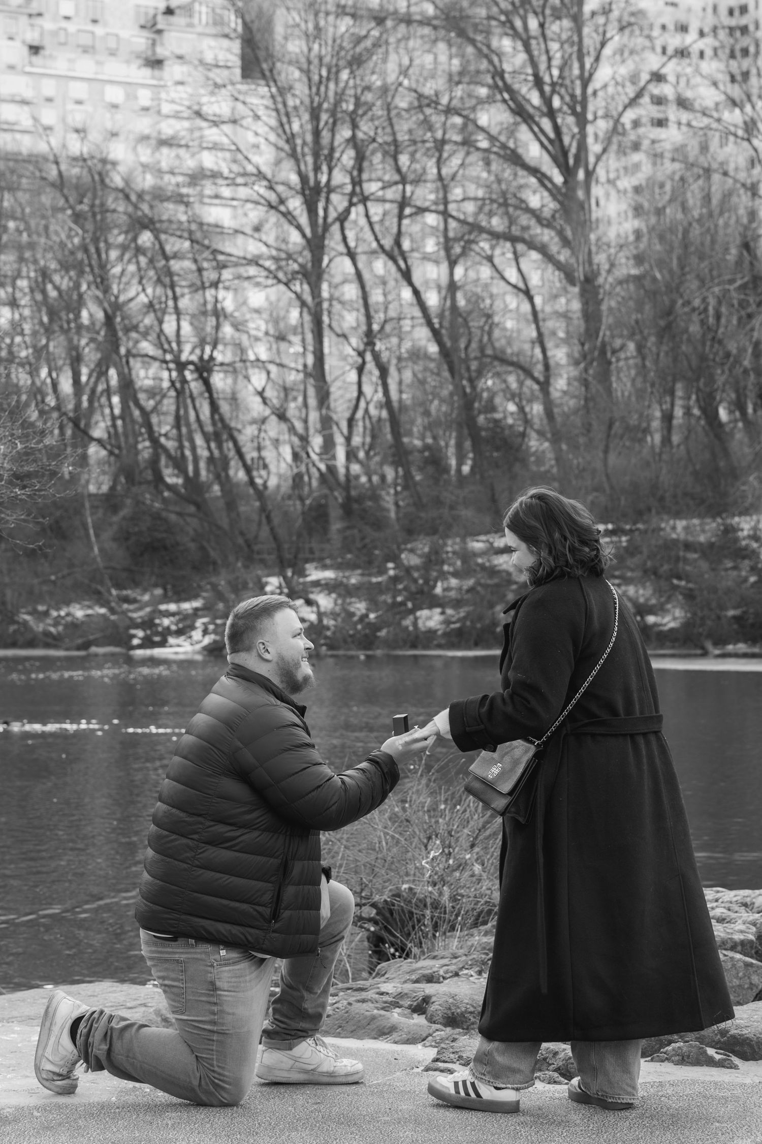 A proposal moment captured in black and white by a lake with bare winter trees and city buildings in the background.
