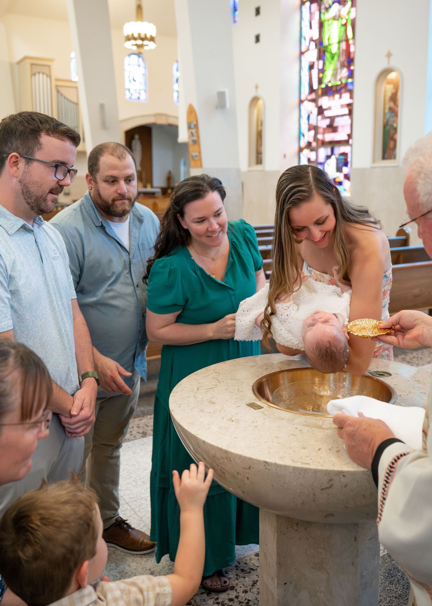 A baptism ceremony takes place around a stone font in a church with stained glass windows.