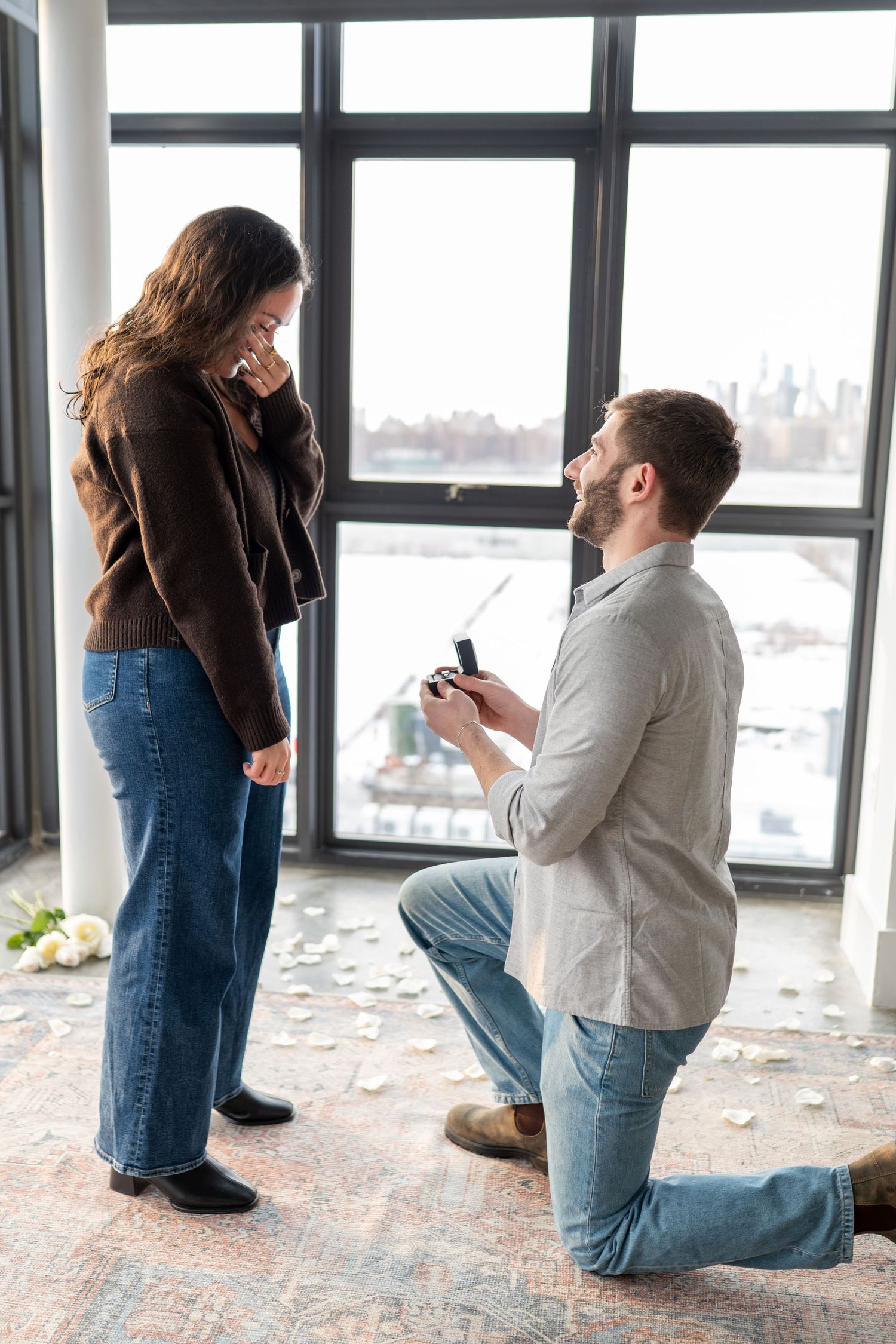 A romantic proposal moment by large windows overlooking a winter cityscape with someone kneeling and presenting a ring.