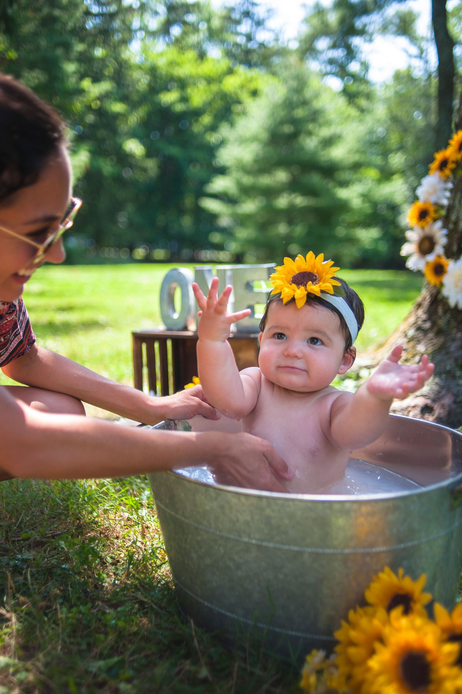 Baby splashing in metal tub during outdoor photoshoot with sunflowers.