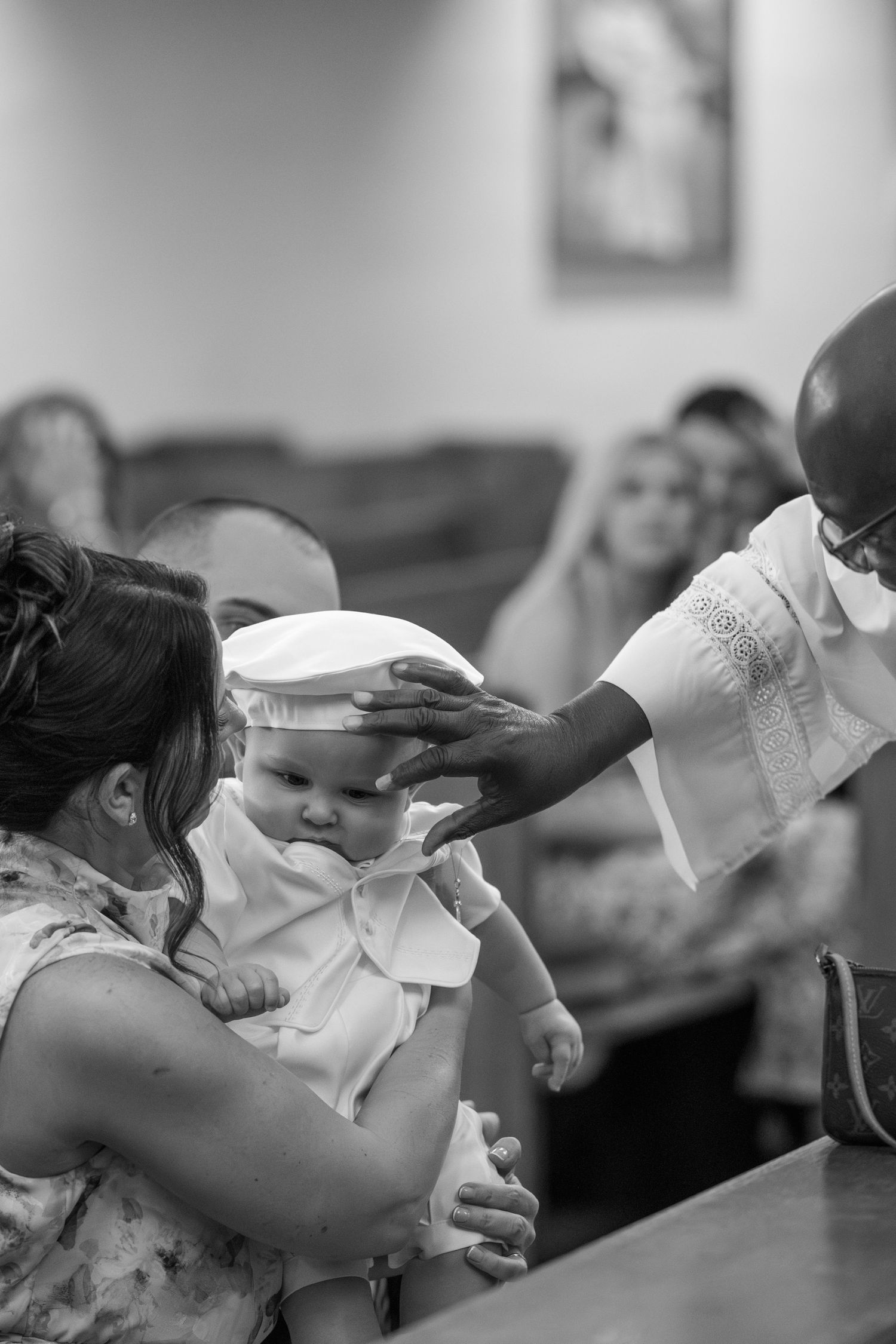 A baby in white clothing receives a blessing during a religious ceremony while being held by someone in a floral dress.