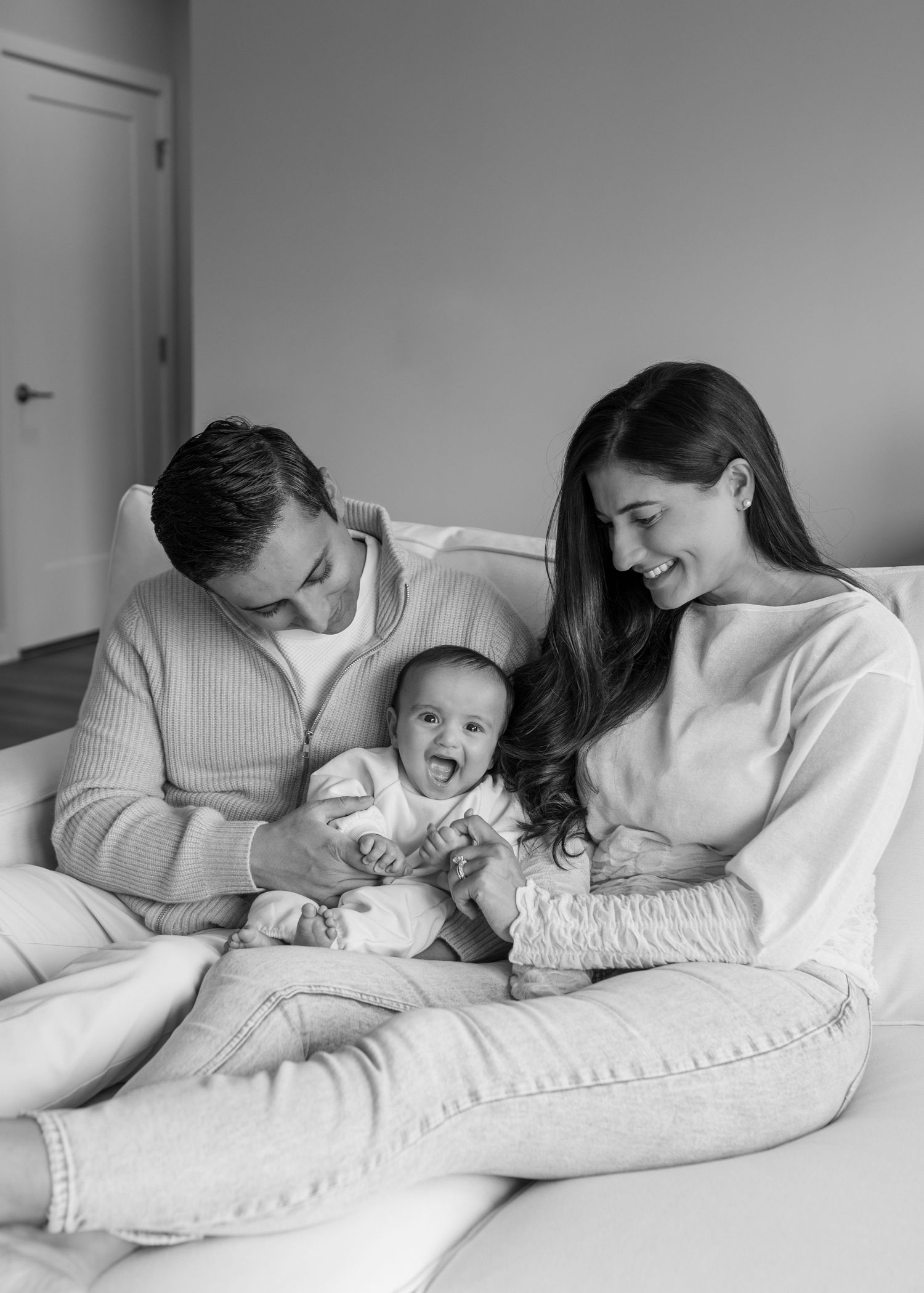 Black and white photo of parents laughing with happy baby on couch in cozy home setting.