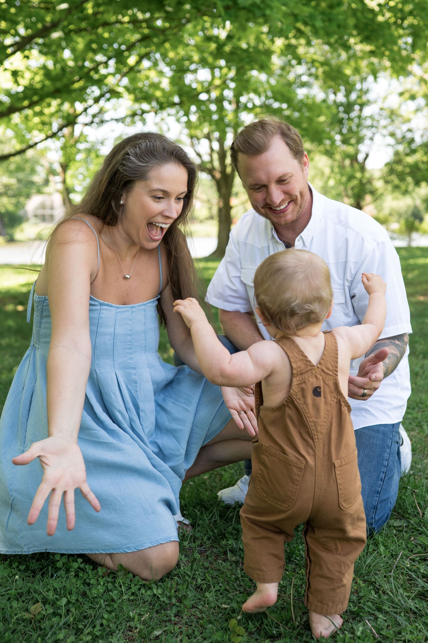 Happy family moment as parents joyfully encourage their baby\u2019s first steps on green grass in a sunny park.