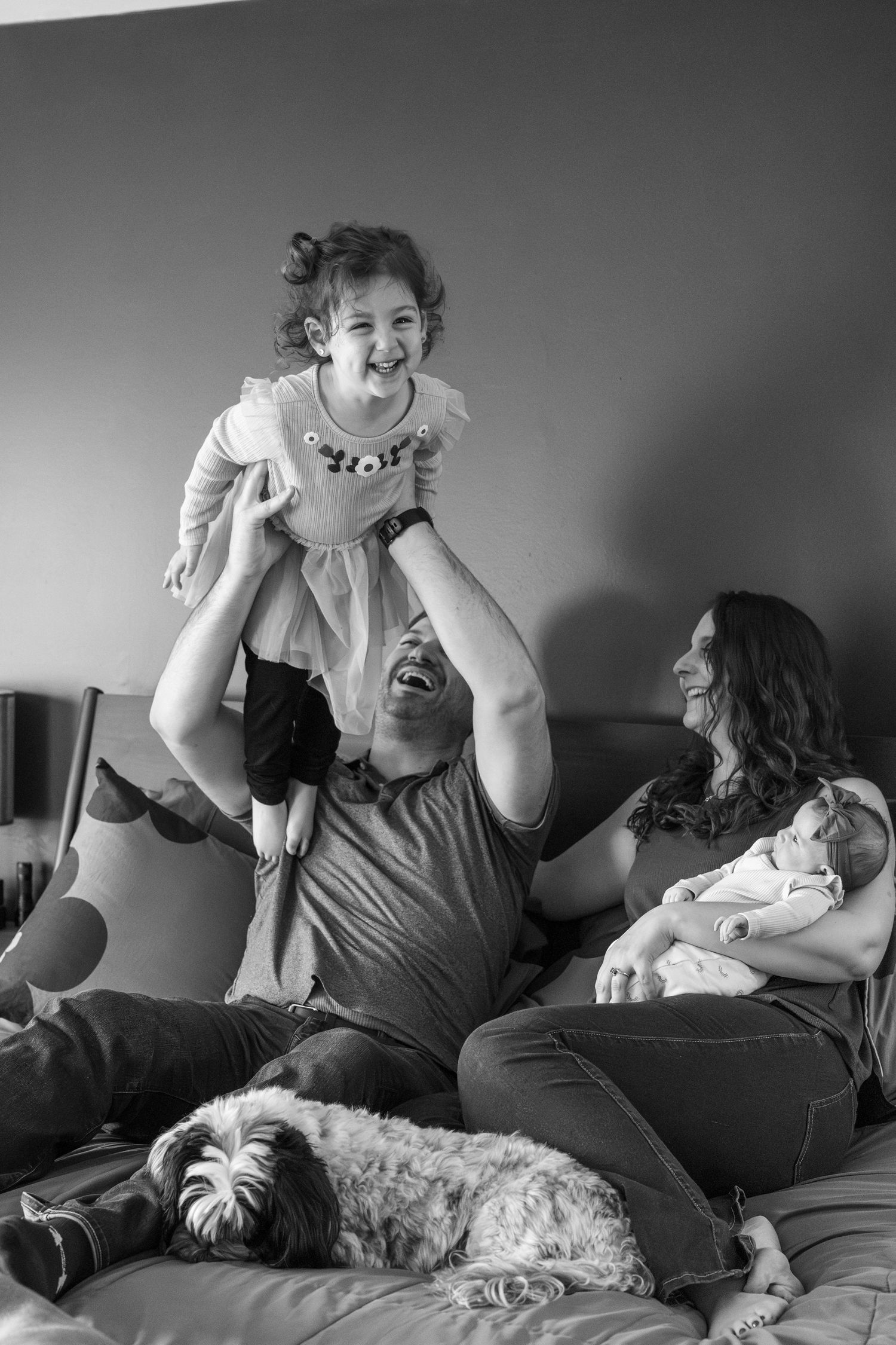 A playful sequence of black and white photos showing people laughing and playing with a small dog on a bed.