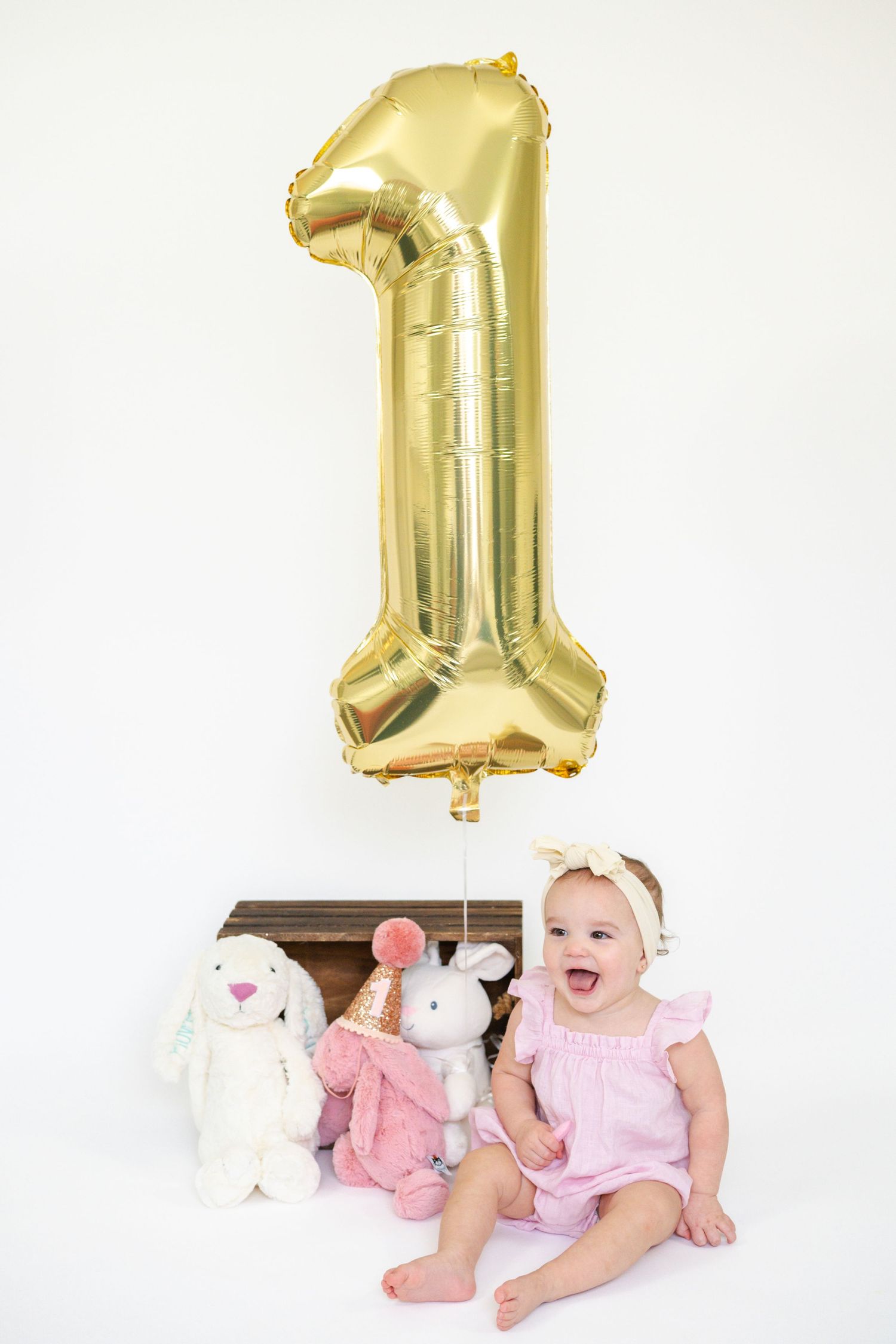 Baby in pink outfit sitting with stuffed animals and gold number one balloon for first birthday celebration.