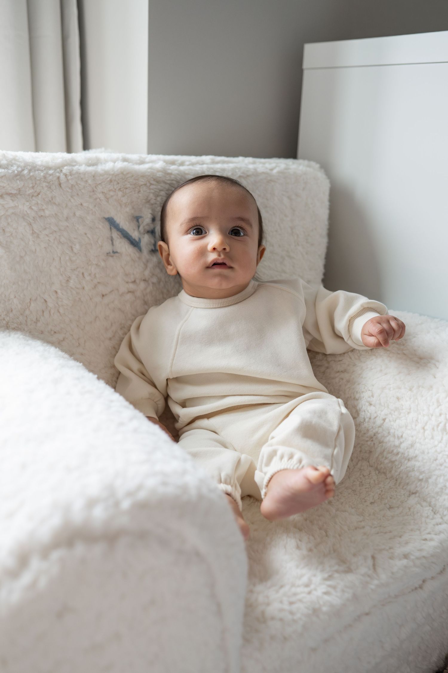 Baby in cream-colored outfit lying on plush white bedding while looking up with curious expression.