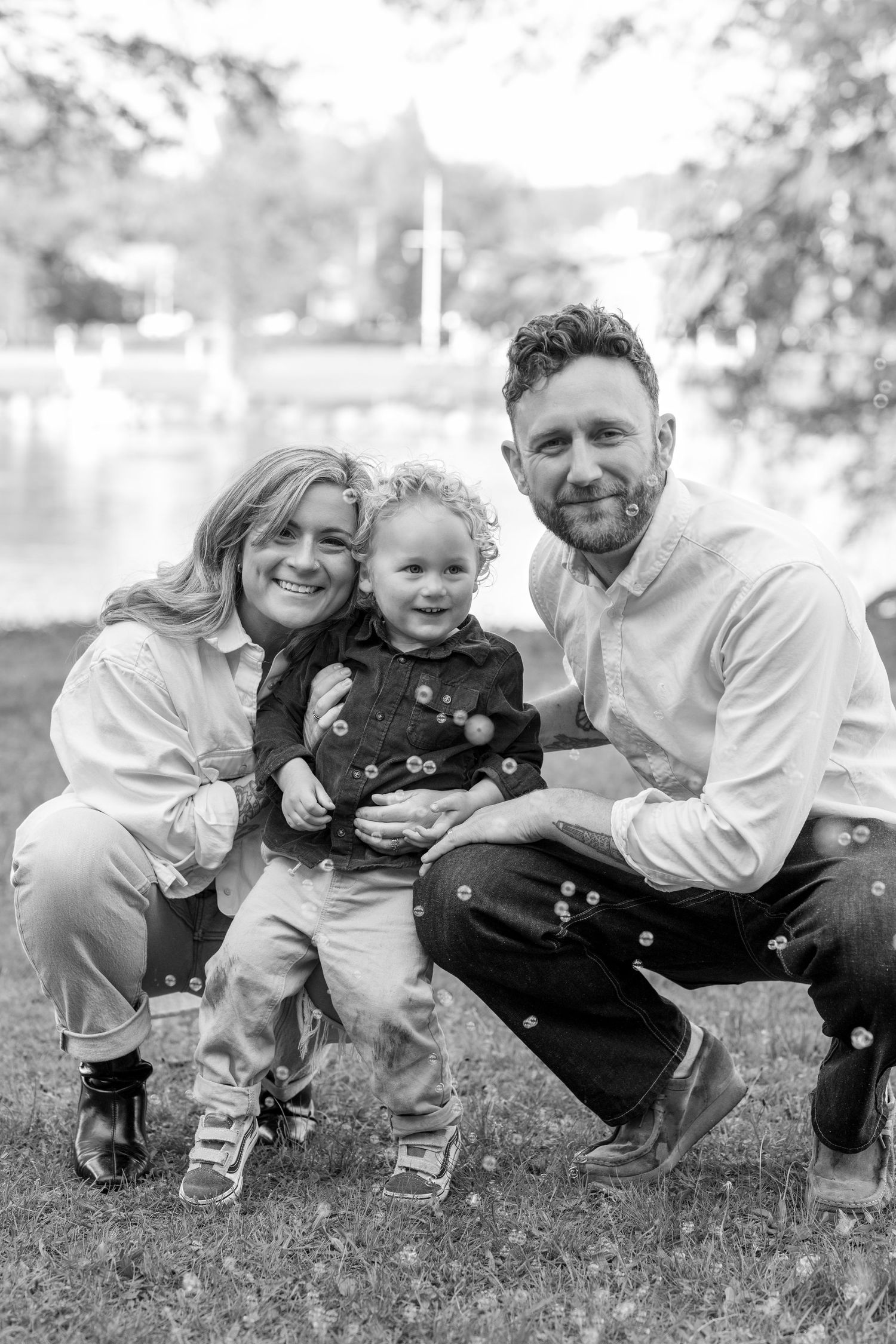 Black and white family portrait of smiling parents crouching with their happy toddler outdoors.