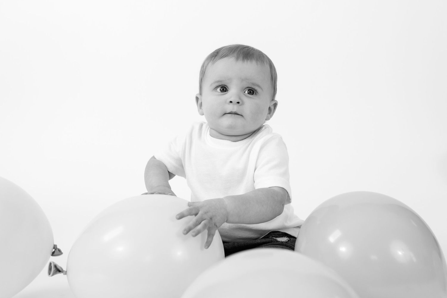Black and white photo of a cute baby in white shirt sitting among balloons on white background.