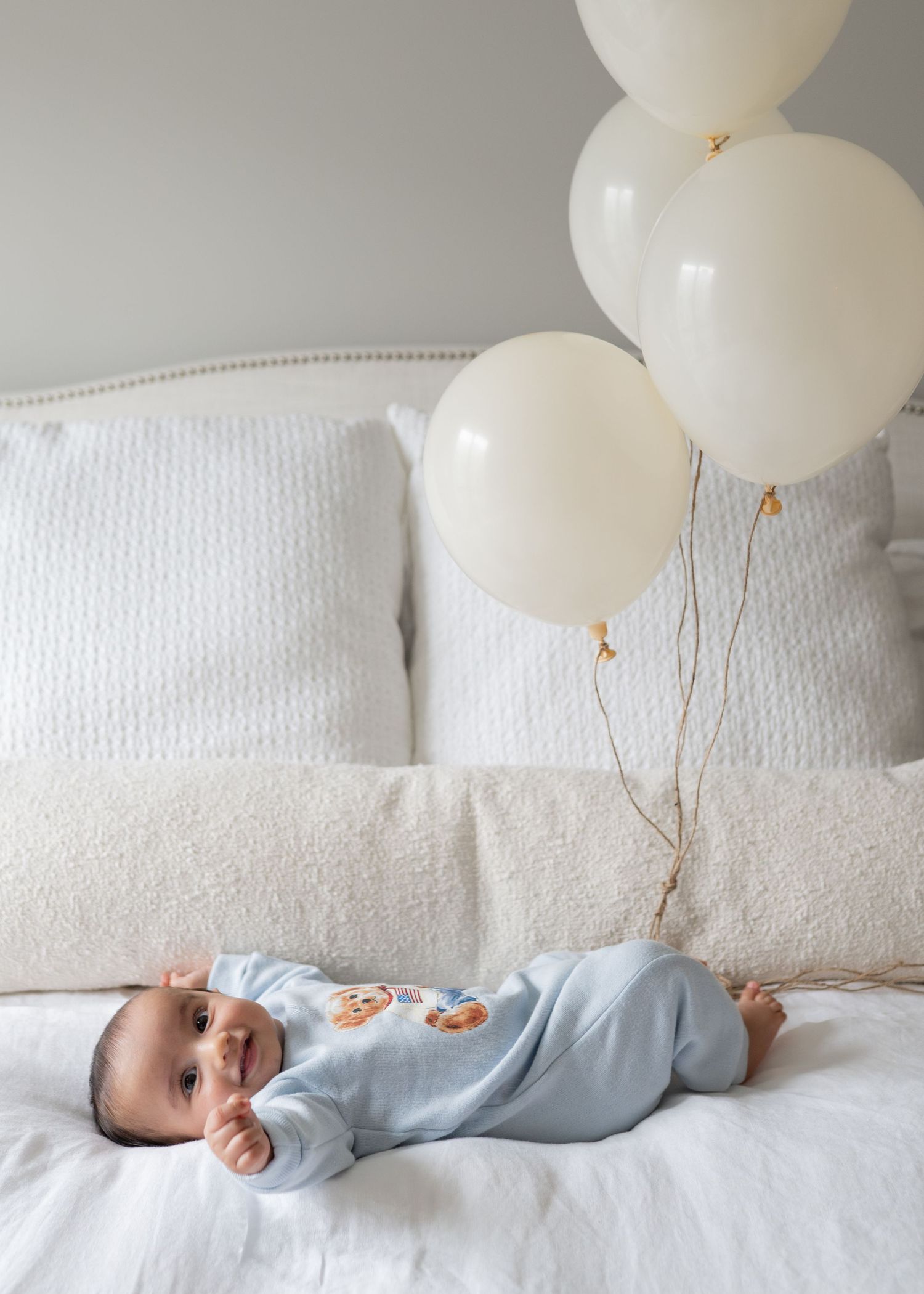 Baby in blue onesie lying on white bed with cream balloons.
