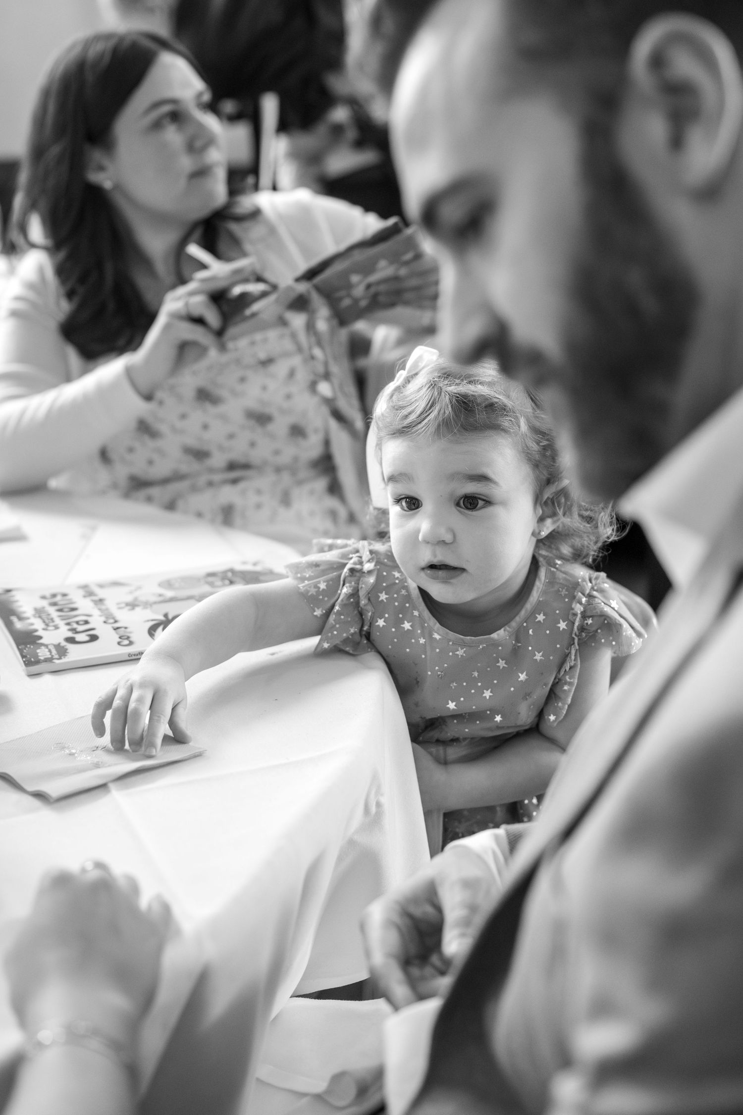 Young child in polka dot dress looking at father while sitting at table with mother in background, black and white family photo.