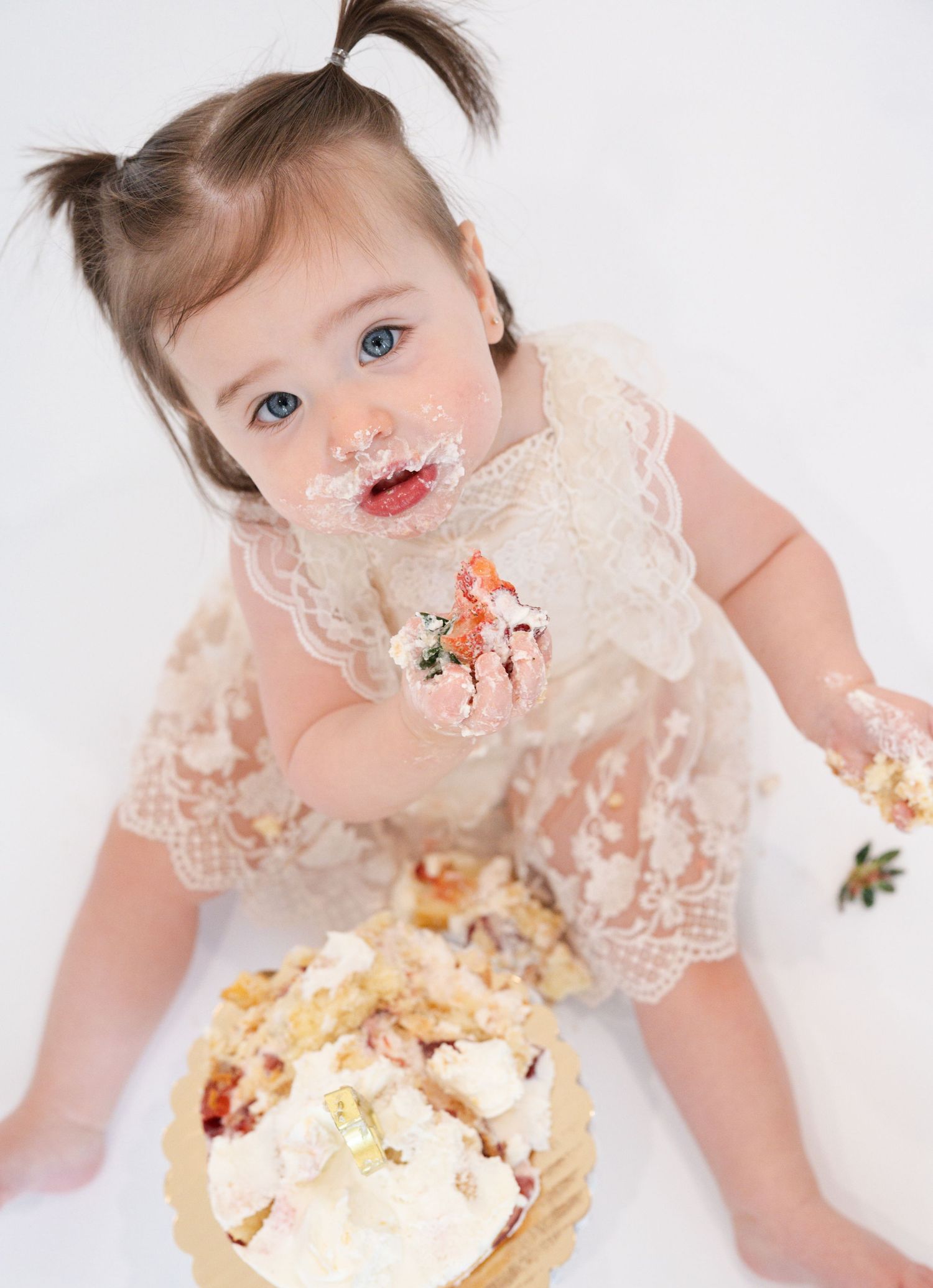 Baby wearing lace dress plays with smashed cake during first birthday photoshoot.