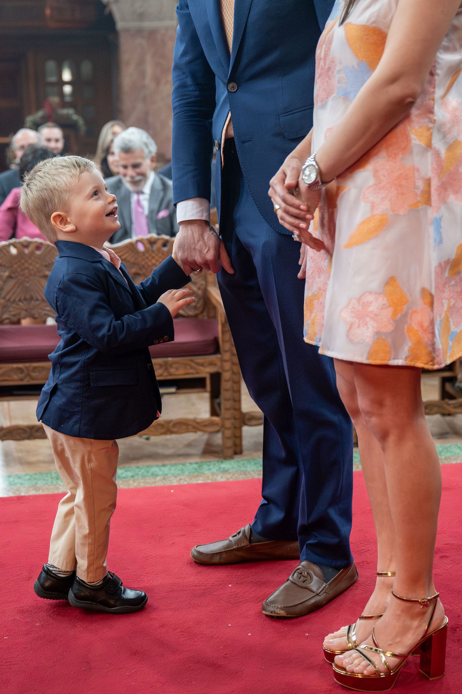 Young boy in navy suit holding adult man's hand on red carpet at formal ceremony.