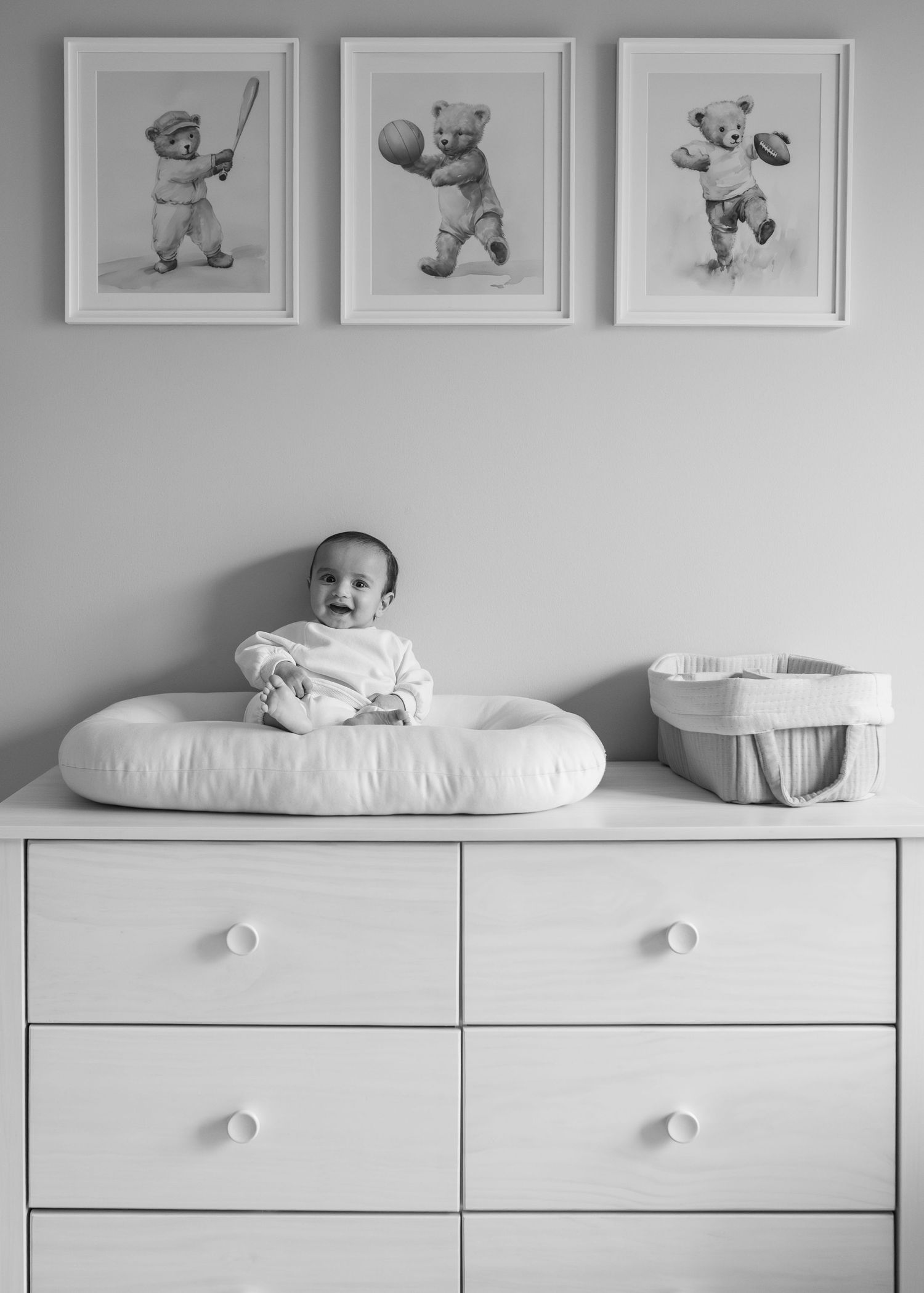 Black and white photo of a smiling baby sitting on a changing pad atop a dresser, with framed teddy bear prints above.