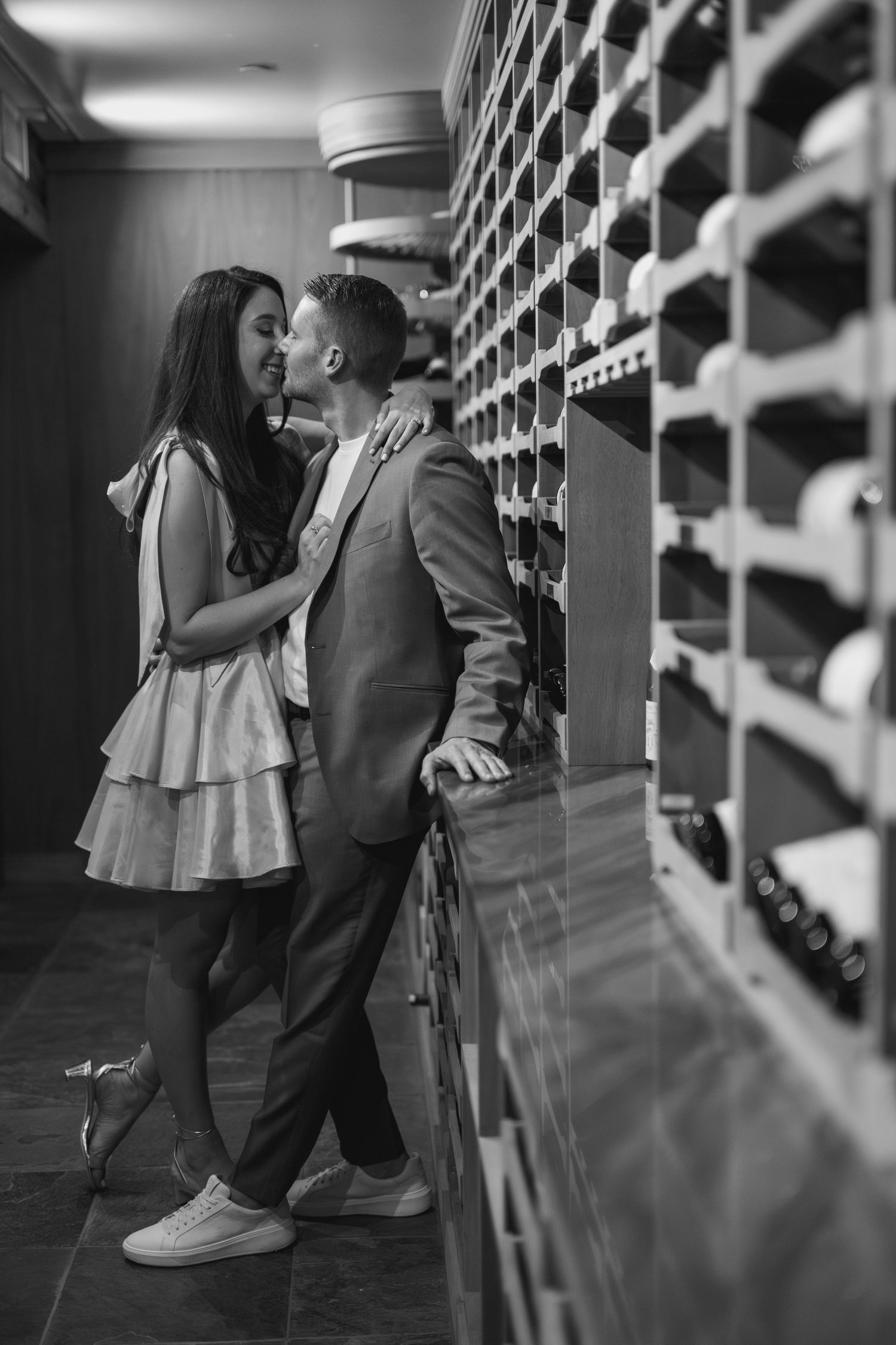 Black and white photo of a romantic couple sharing an intimate moment in an elegant wine cellar with bottle-lined shelves.