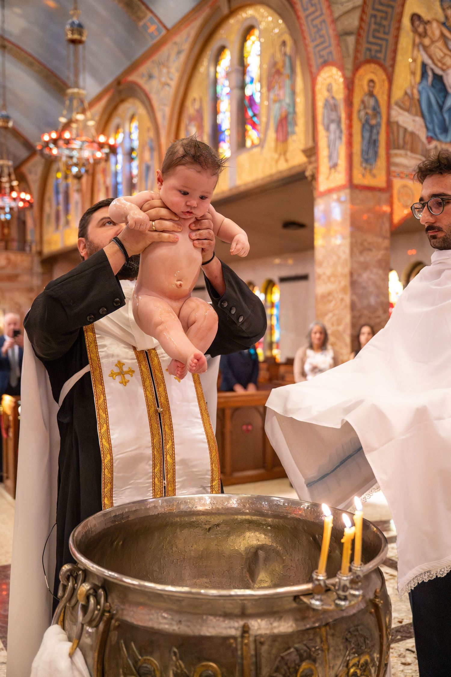 A baptismal ceremony takes place in an ornate Orthodox church with golden decorations and stained glass windows.