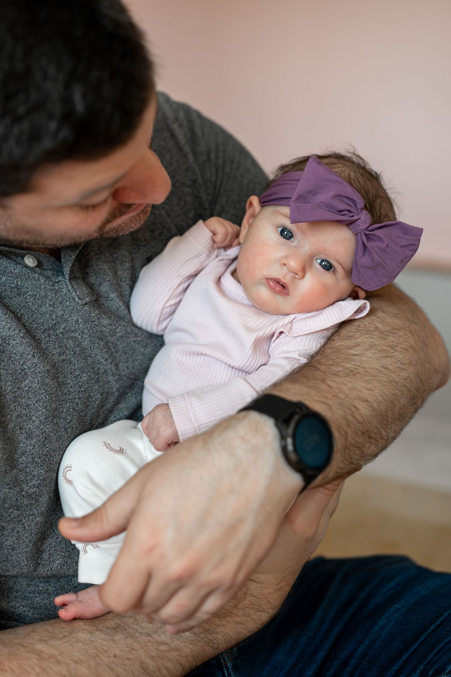 A baby wearing a purple bow and white outfit rests on someone's arm.