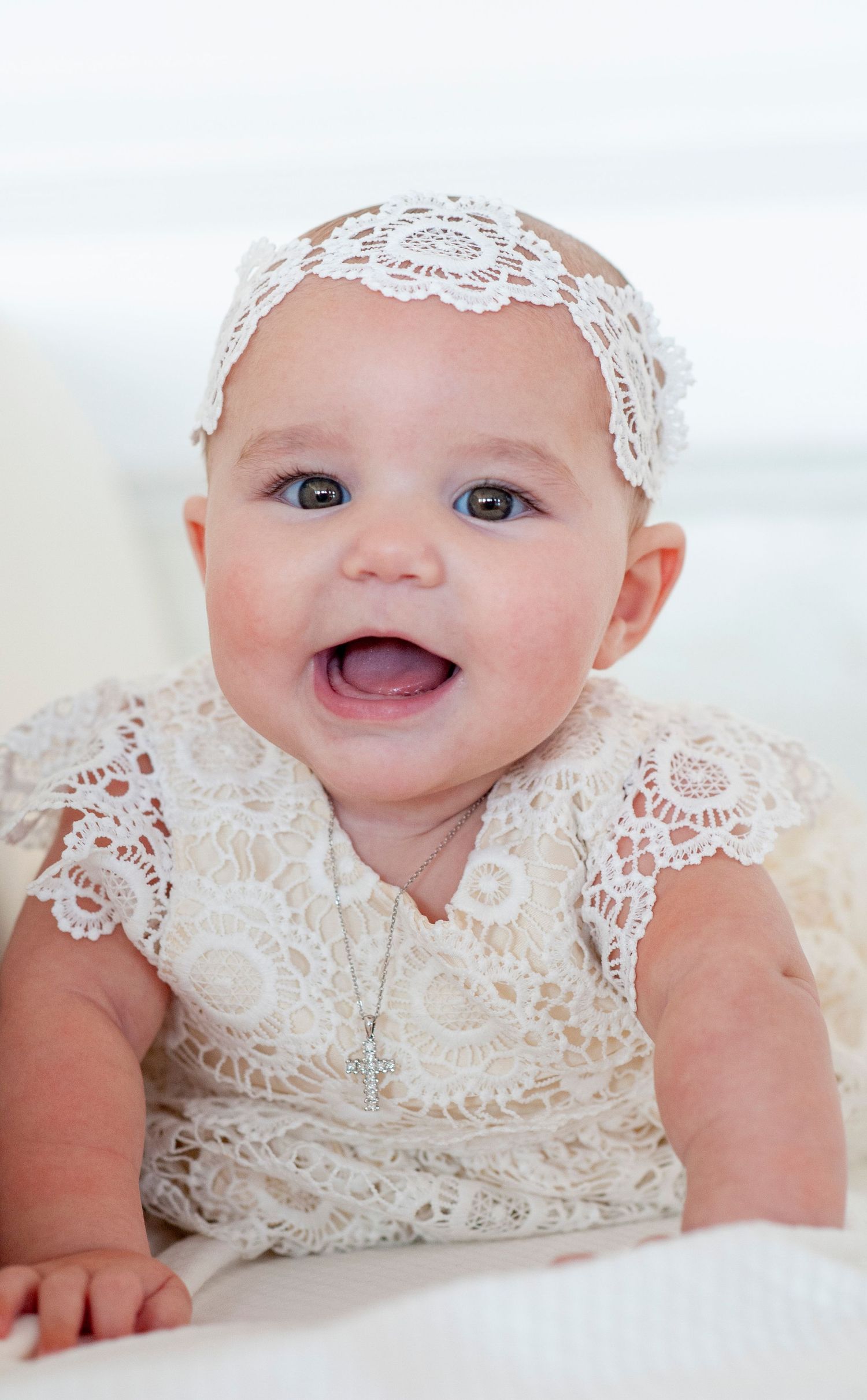 A baby wearing delicate white lace clothing smiles while lying down.