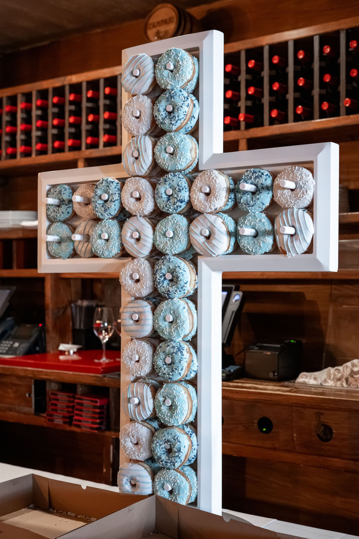 Cross-shaped display made of stacked blue and white decorative donuts against wooden wine rack backdrop.