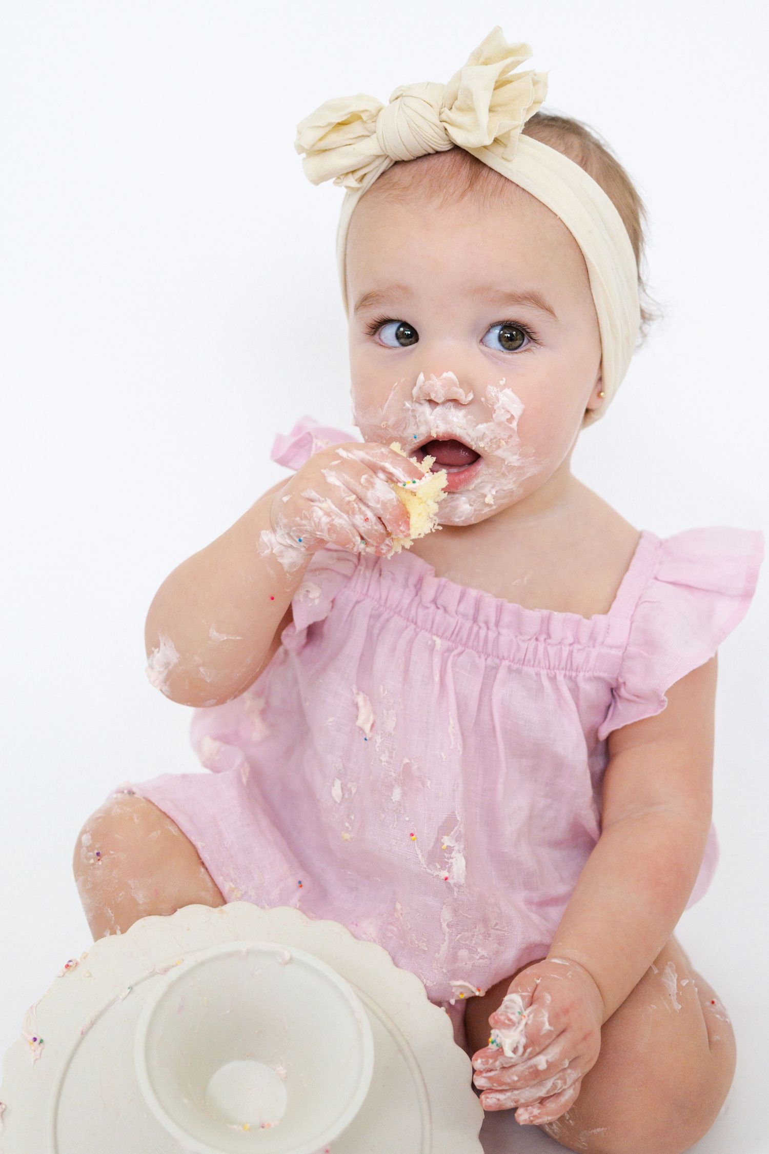 Baby in pink dress with bow headband getting messy with birthday cake during photoshoot.