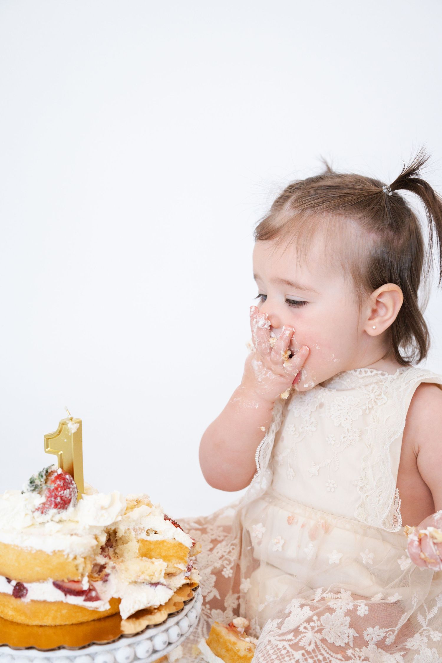 Baby in lace dress enjoying their first birthday cake smash photoshoot against white background.