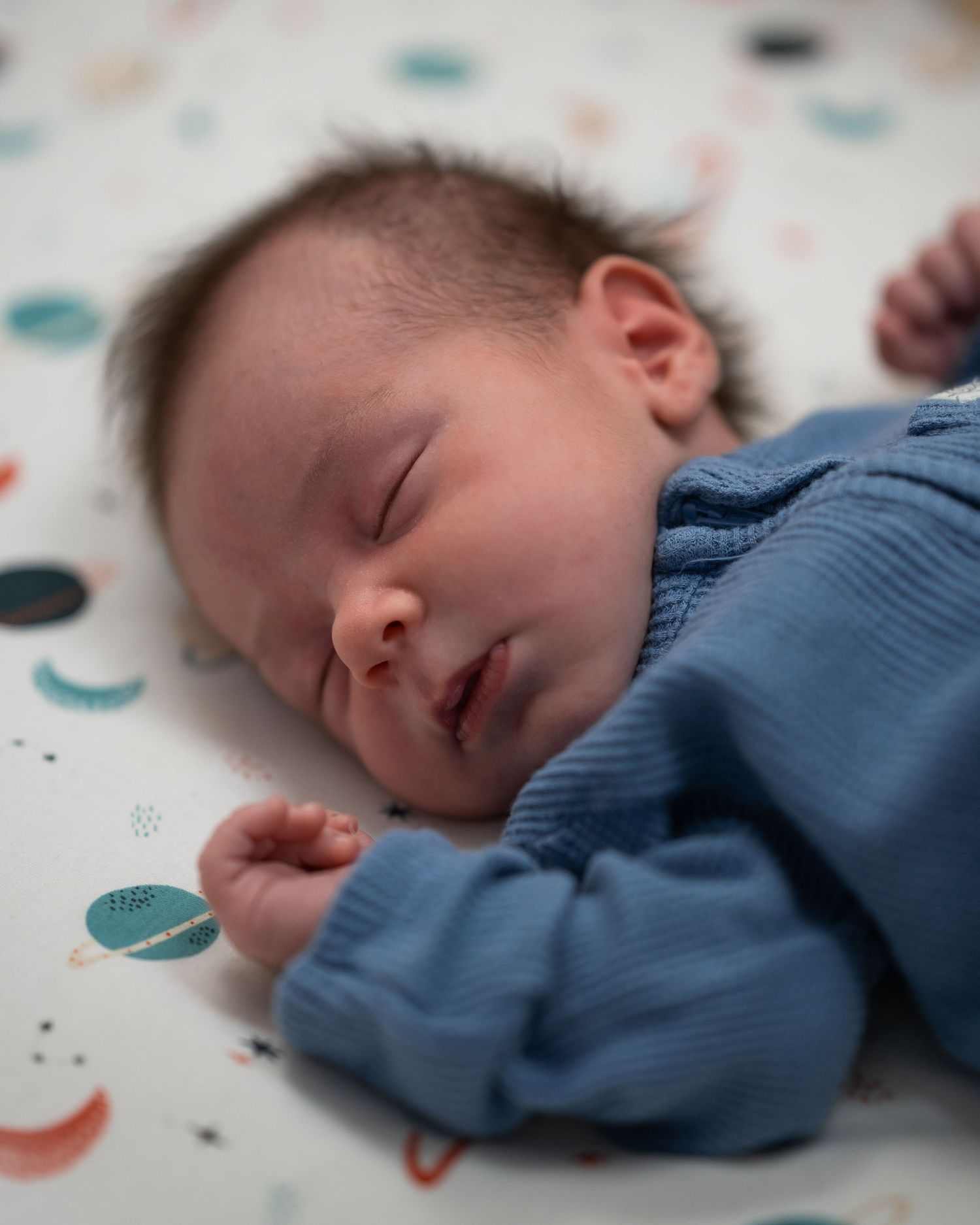 Sleeping baby in blue outfit rests on patterned bedding.