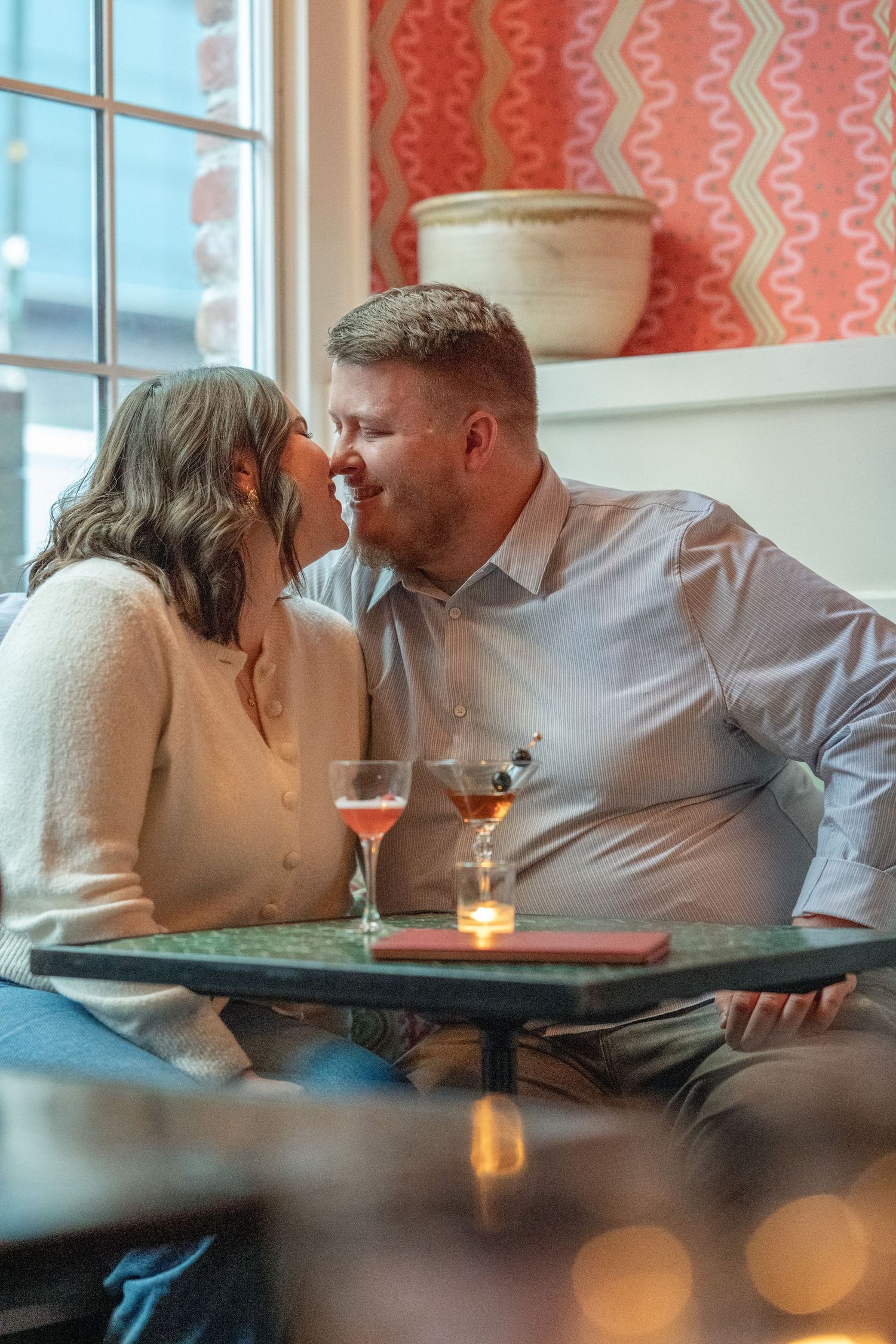 A romantic couple shares an intimate moment at a restaurant table with candlelight and pink decor.